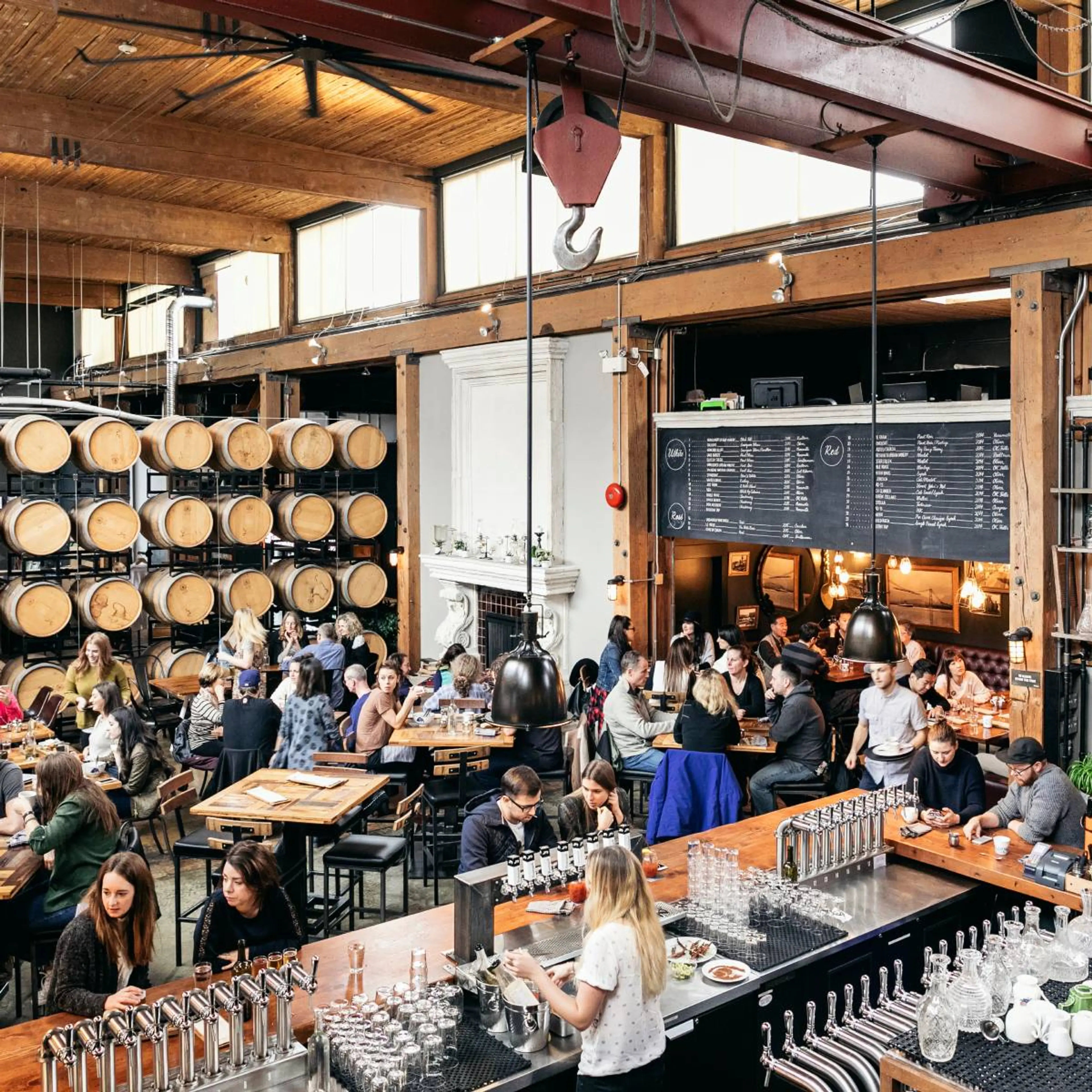 Busy brewery taproom with wooden barrels, patrons sitting at tables and bar, and large menu boards on the wall.