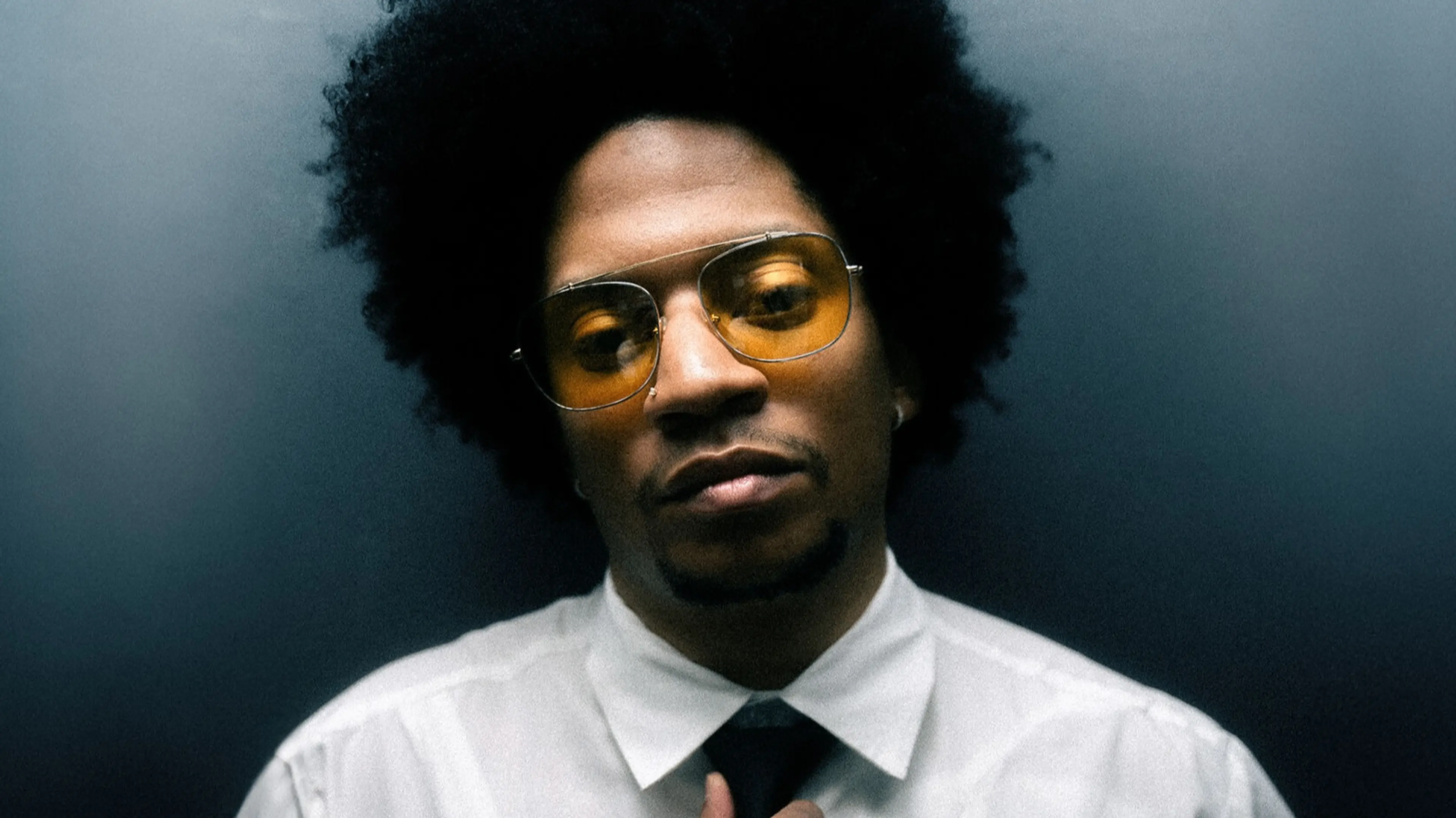 Person with afro hairstyle wearing yellow-tinted glasses, white shirt, and black tie against a dark background.