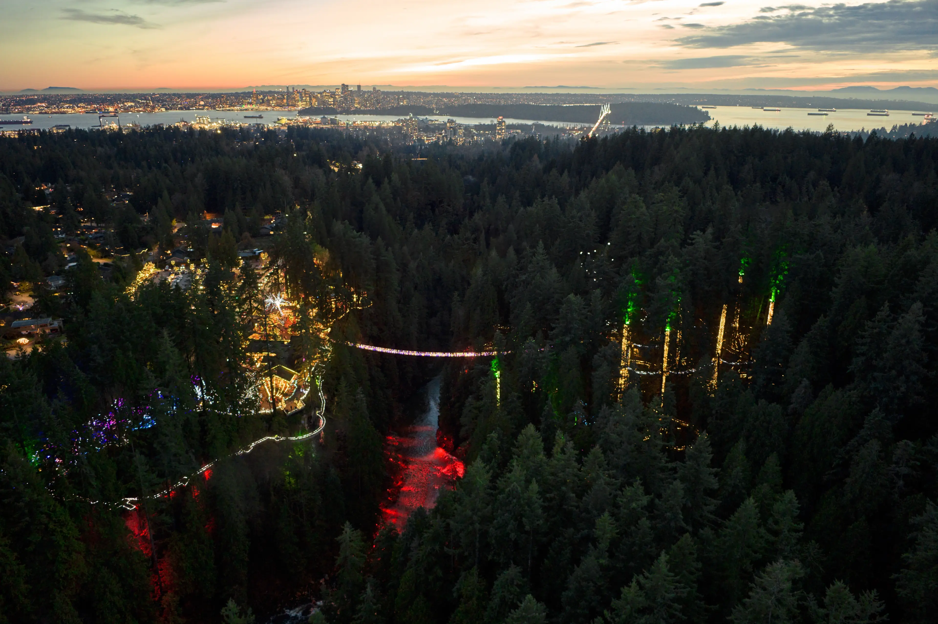 Aerial image of the Capilano Suspension Bridge lit up with Christmas lights and the Vancouver city skyline in the background.
