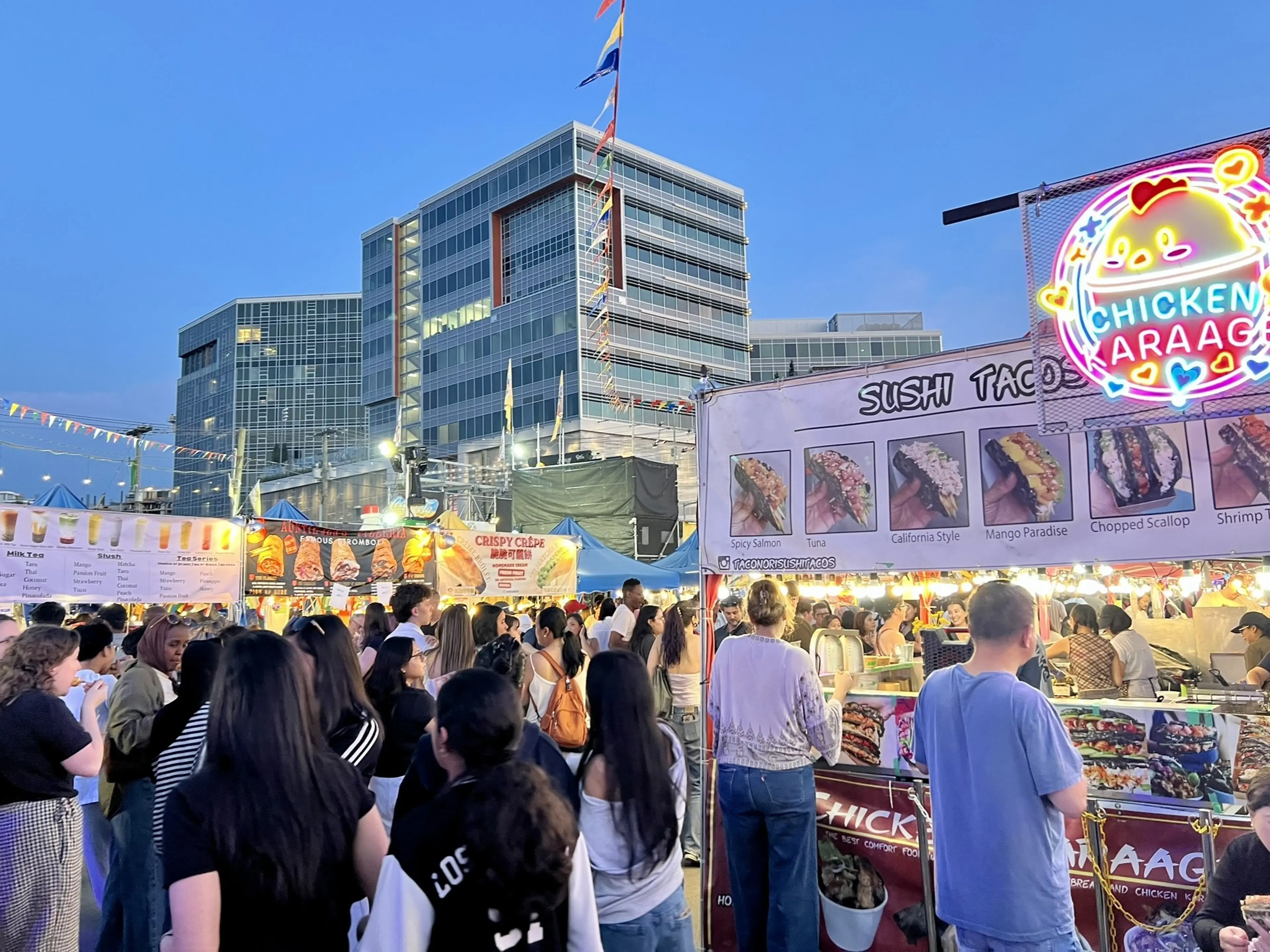 Crowd at an outdoor food market with sushi taco and chicken karaage stalls under evening sky and modern buildings in the background.