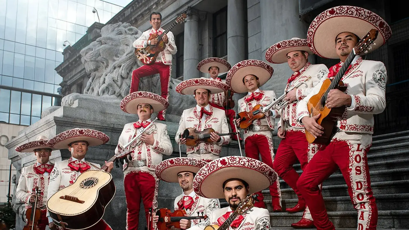 A mariachi band of eleven people wearing white charro suits with red embroidery and large sombreros, holding guitars, violins, and trumpets, posing on stone steps with a lion statue in the background.