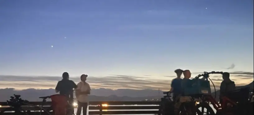 Four people standing near bicycles at dusk with a bench and a scenic sky in the background.