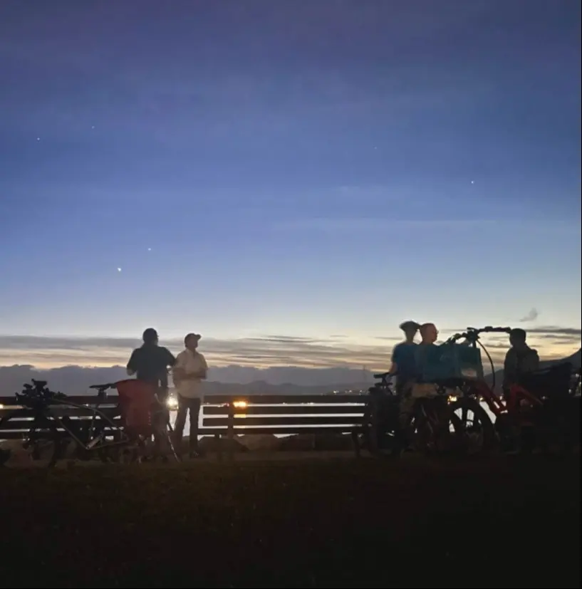 Four people standing near bicycles at dusk with a bench and a scenic sky in the background.
