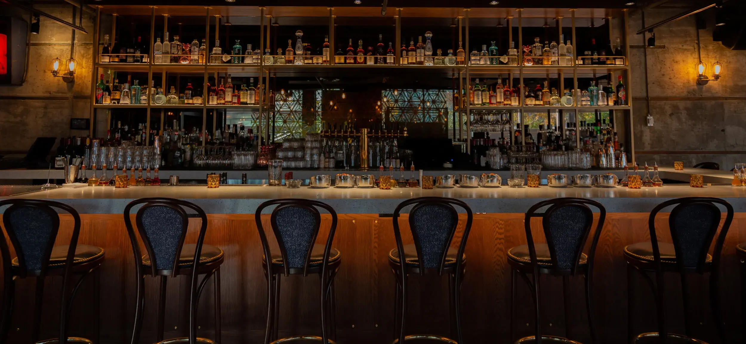 Empty bar counter with seven high chairs and shelves stocked with bottles and glasses in warm lighting