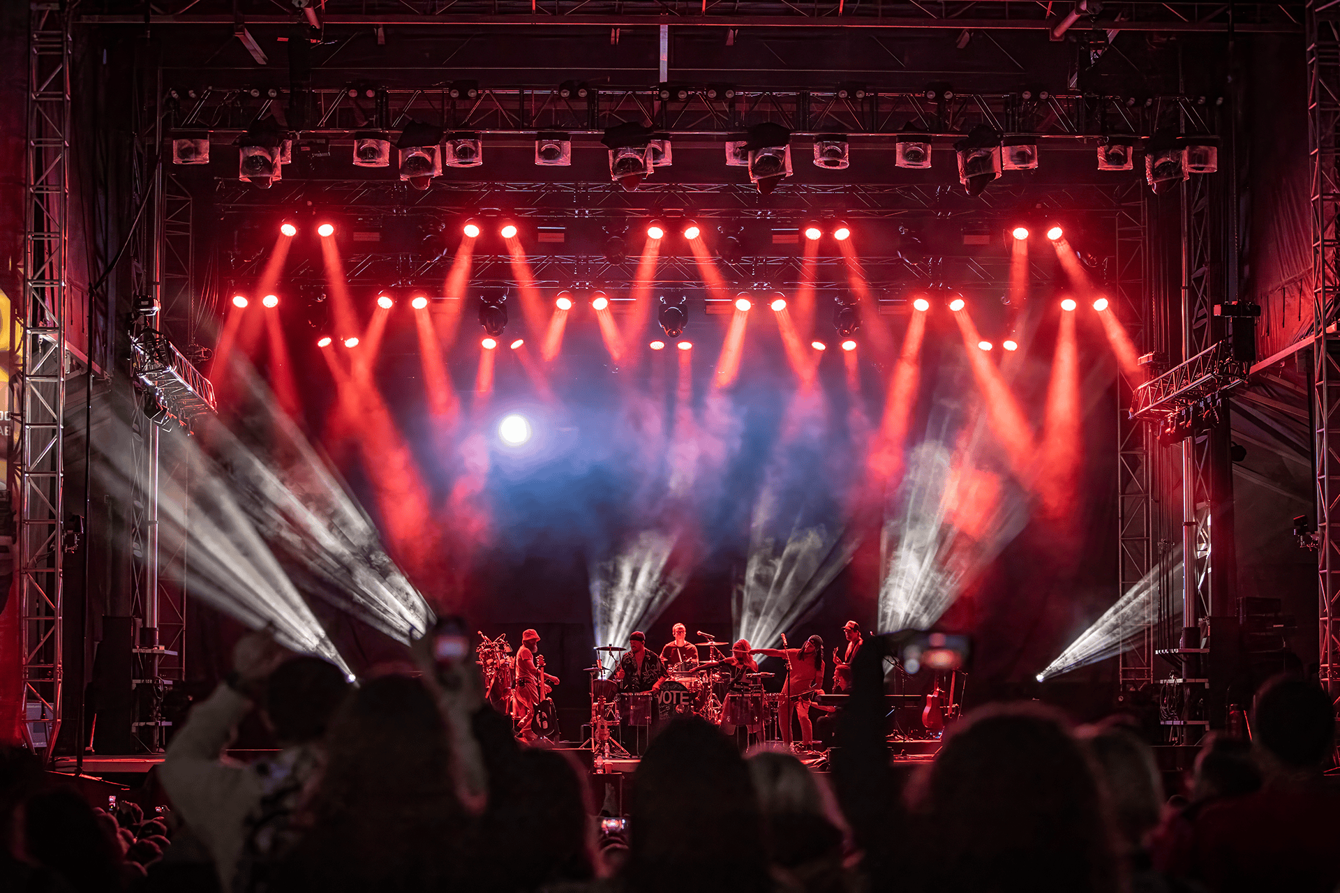 Band performing on stage with red and white spotlights and audience in foreground capturing the moment on phones.