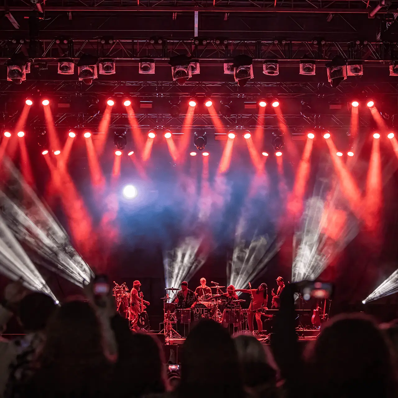 Band performing on stage with red and white spotlights and audience in foreground capturing the moment on phones.