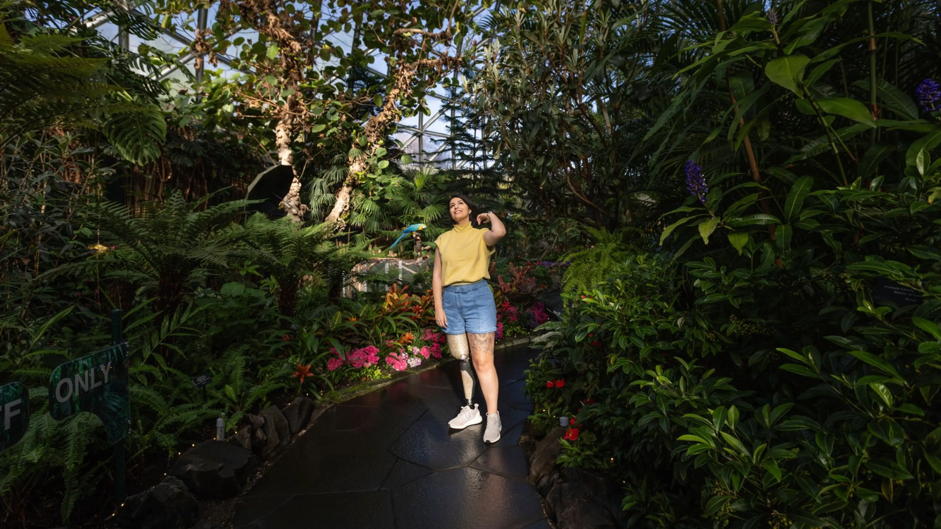 A woman with a prosthetic leg stands on a smooth, paved path surrounded by vibrant tropical plants and flowers inside Bloedel Conservatory.