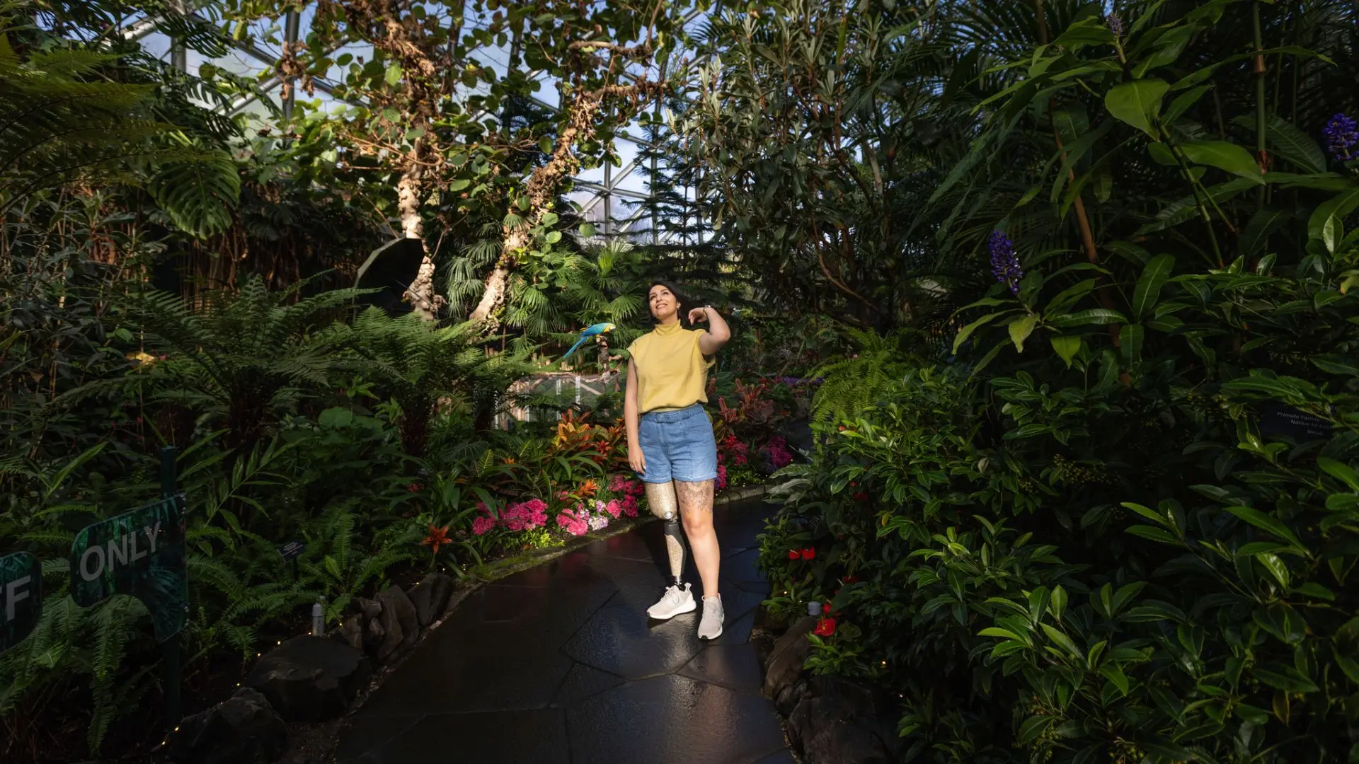 A woman with a prosthetic leg stands on a smooth, paved path surrounded by vibrant tropical plants and flowers inside Bloedel Conservatory.