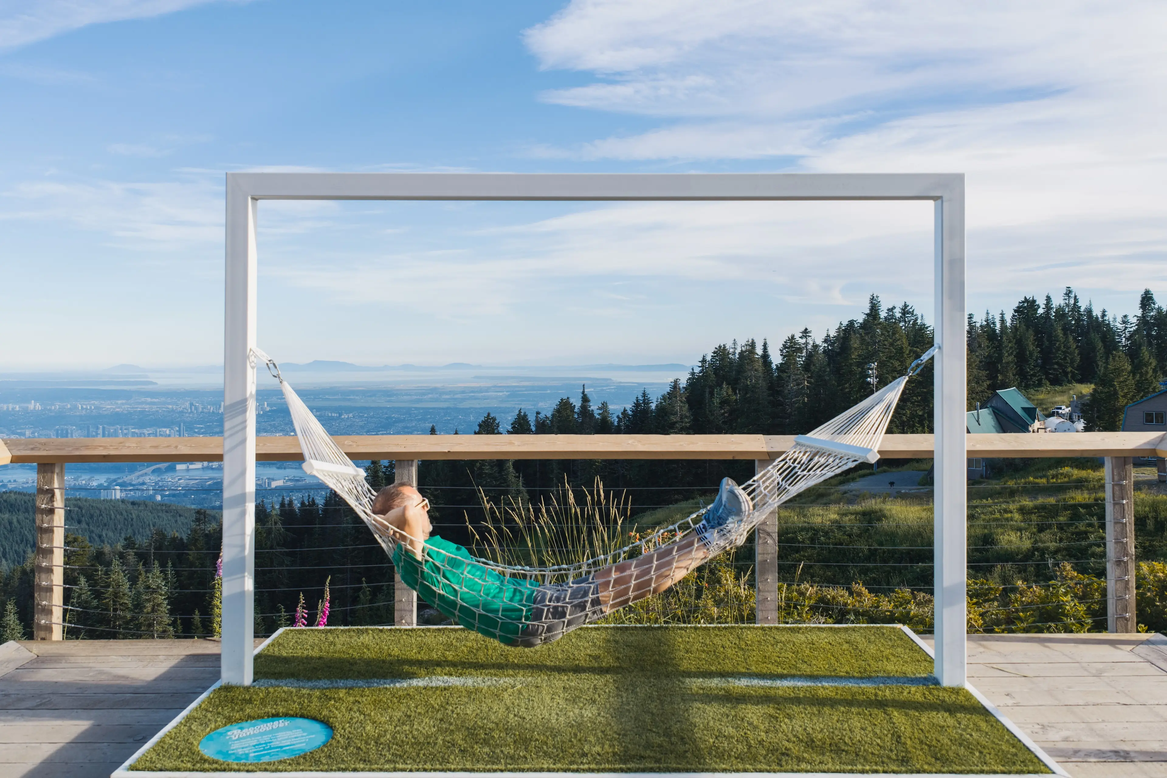 Person relaxing in a hammock framed by a rectangular structure with a scenic mountain and city view in the background.
