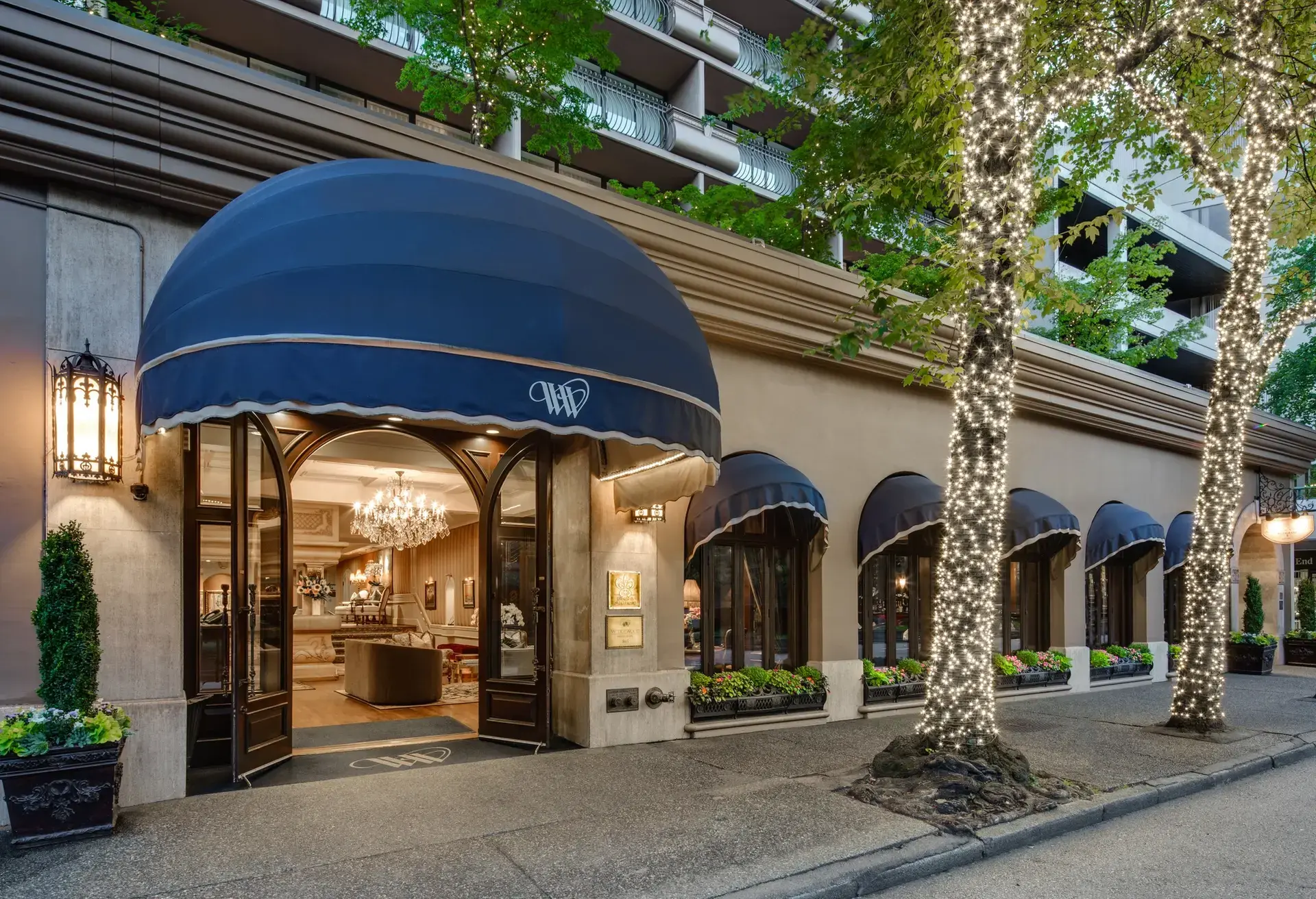 Building entrance with large blue awning, open double doors, chandelier inside, and trees wrapped in white string lights