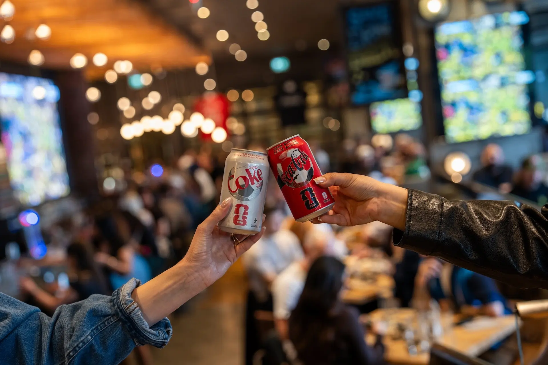 Two people clinking cans of Diet Coke and Coca-Cola in a busy restaurant or bar with blurred background.