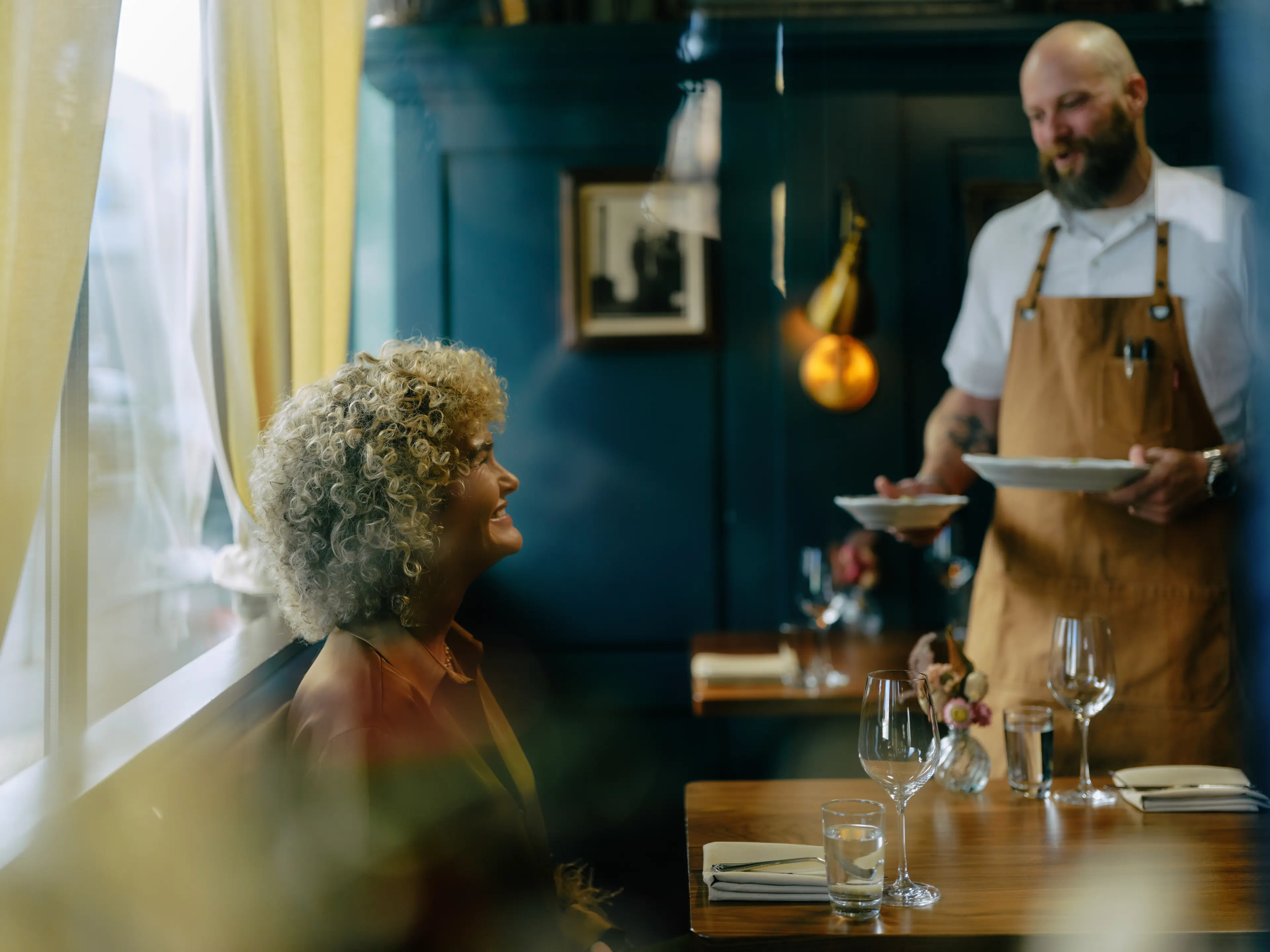 Curly-haired person smiling at a bearded waiter in an apron serving dishes in the restaurant St. Lawrence in Vancouver.