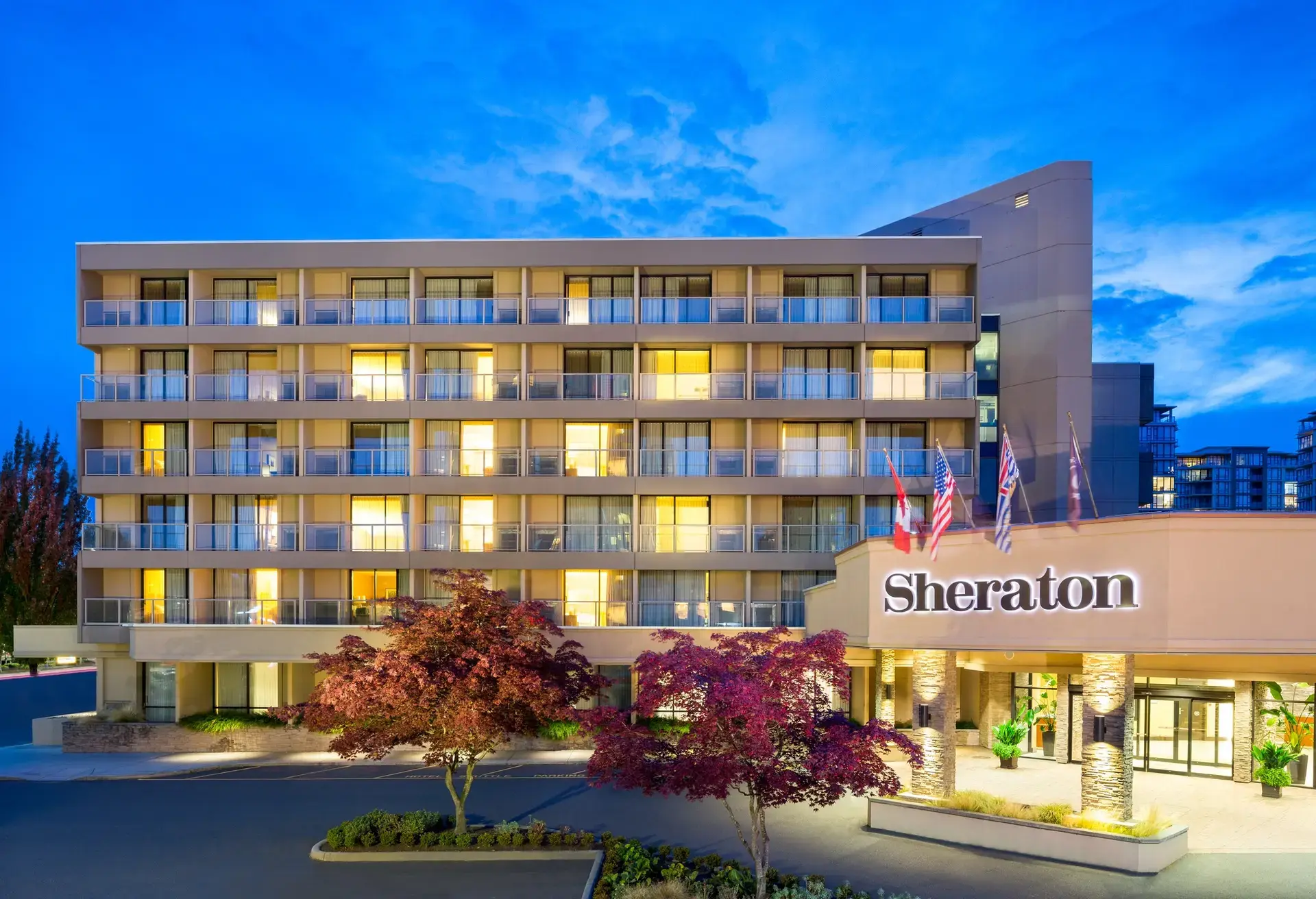 Exterior of a Sheraton hotel building at dusk with lit windows and flags above the entrance.