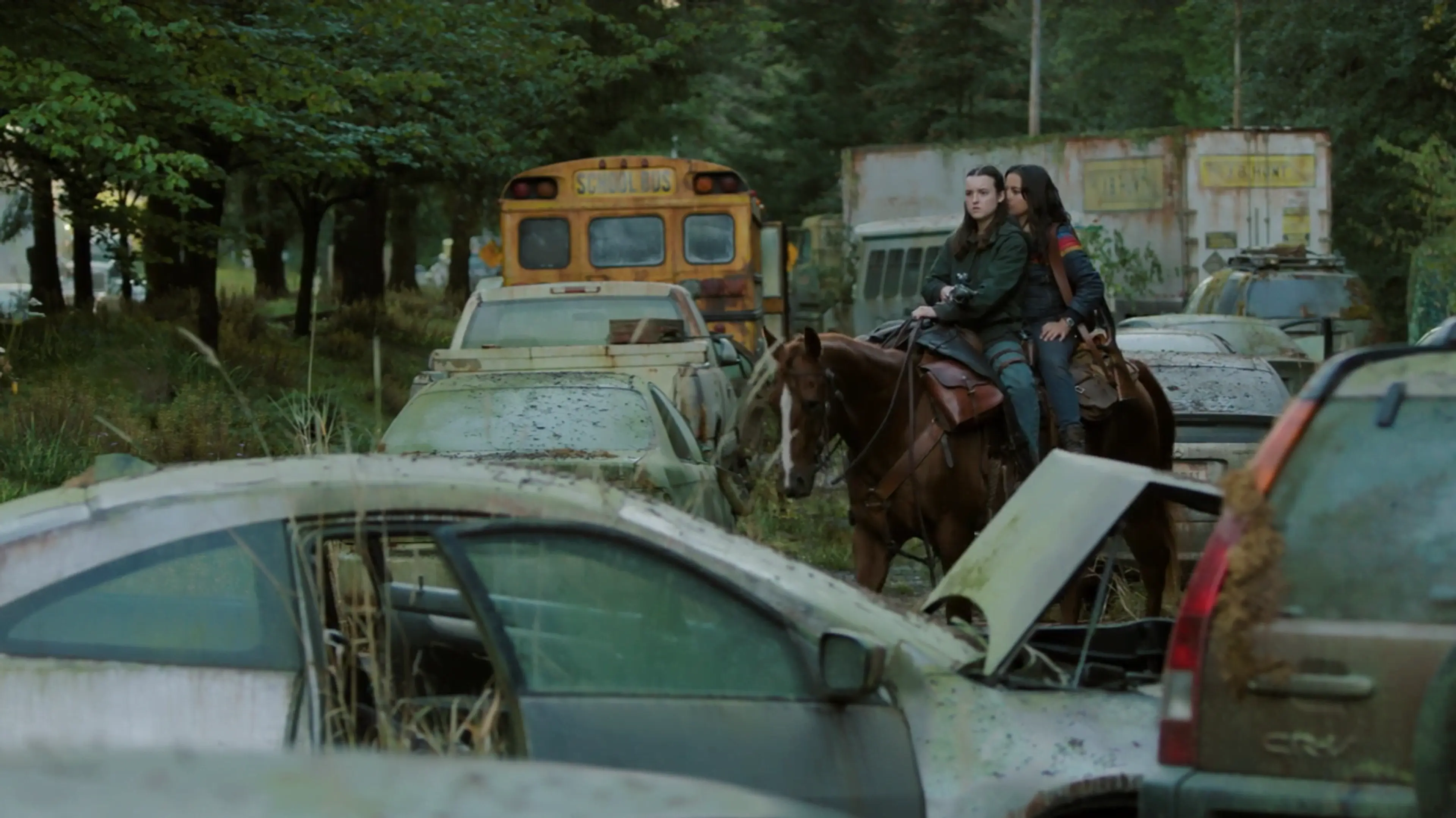 Two people riding a brown horse through an overgrown junkyard filled with abandoned cars and a school bus.