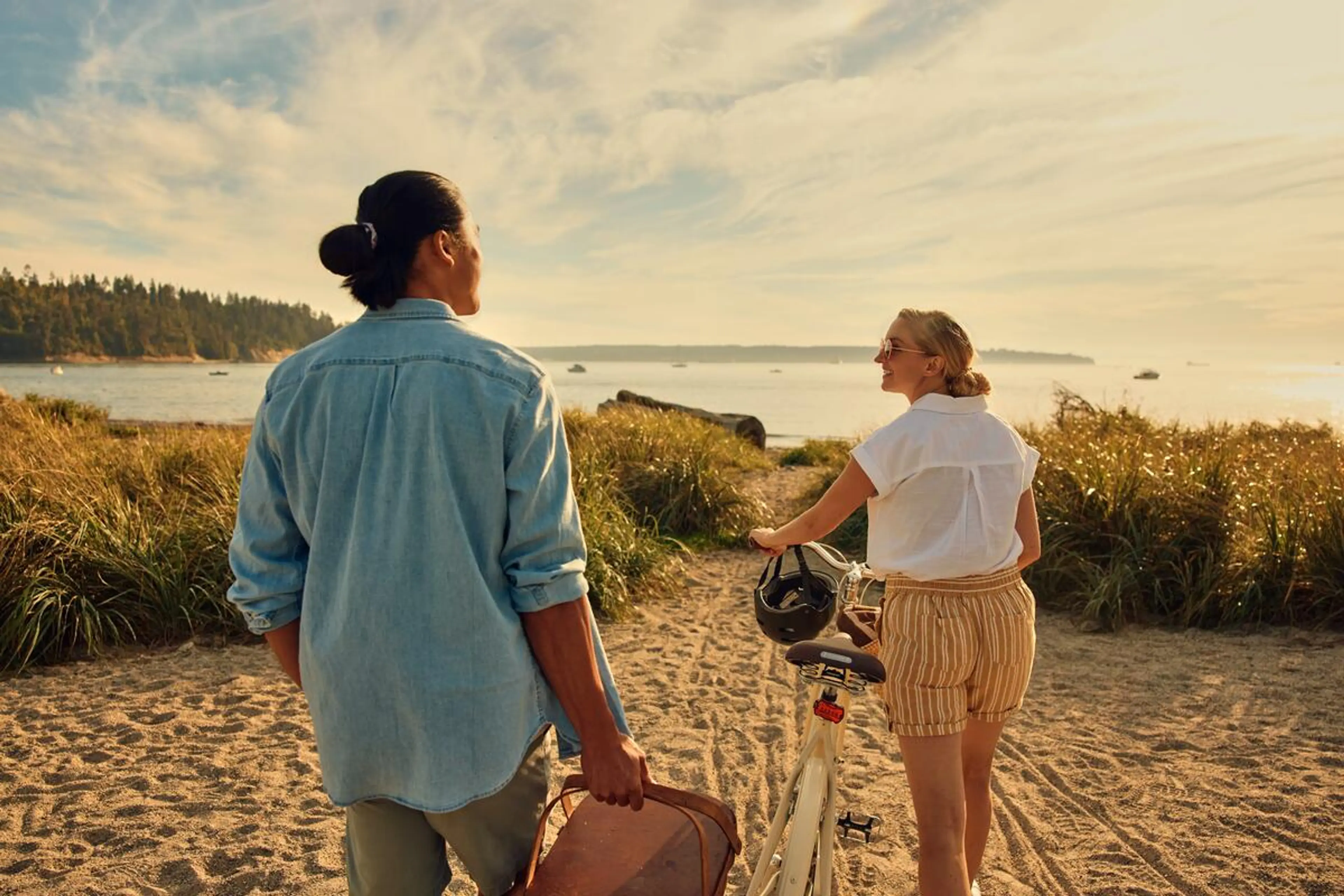 Two people walking on a sandy path toward a body of water, one holding a suitcase and the other pushing a bicycle with a helmet.