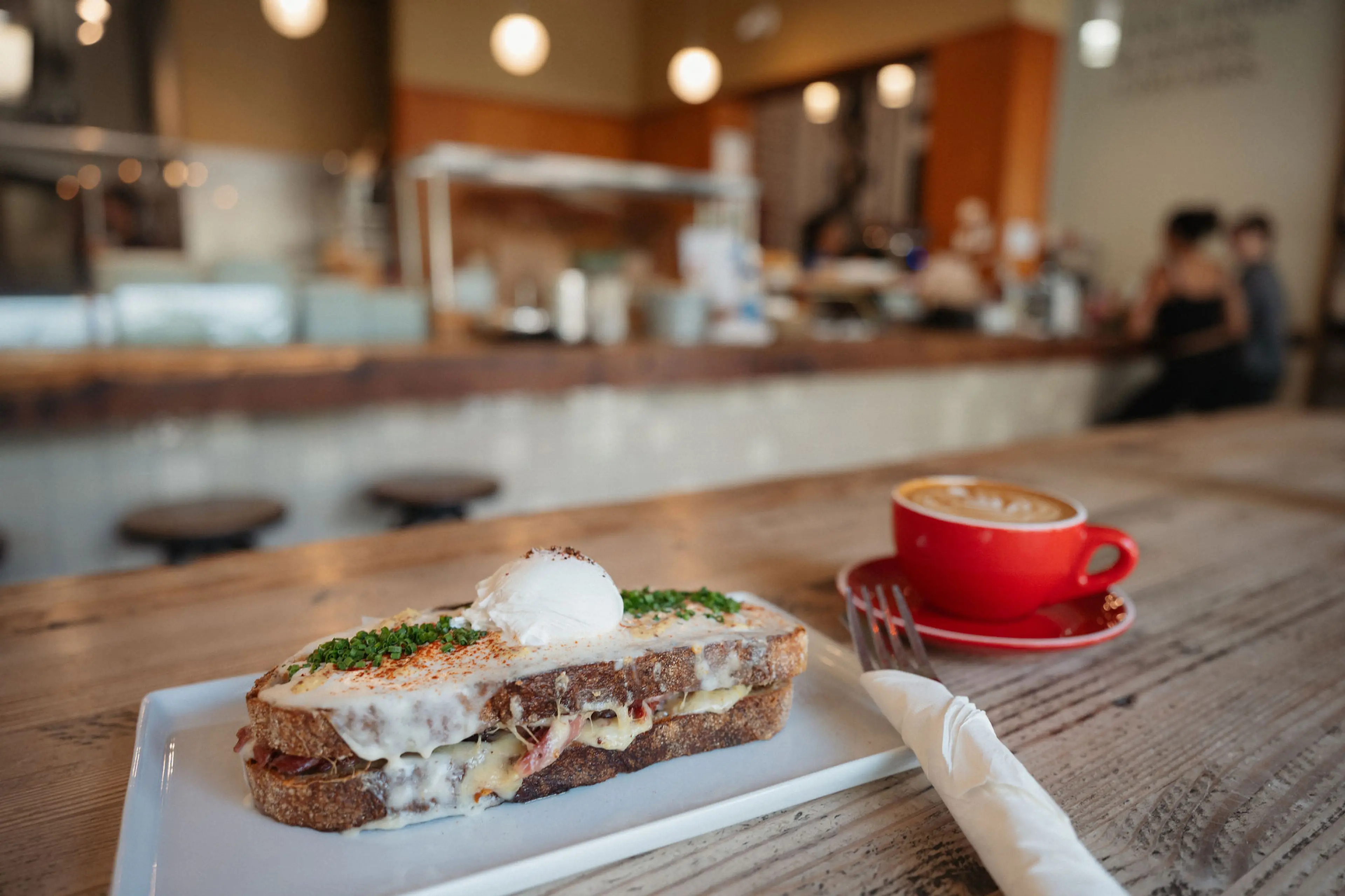 Grilled sandwich with melted cheese and herbs topped with a poached egg on a white plate, next to a red cup of coffee on a wooden table in a cafe.