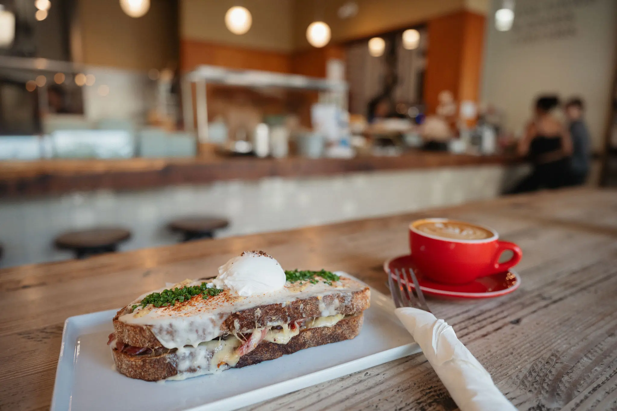 Grilled sandwich with melted cheese and herbs topped with a poached egg on a white plate, next to a red cup of coffee on a wooden table in a cafe.