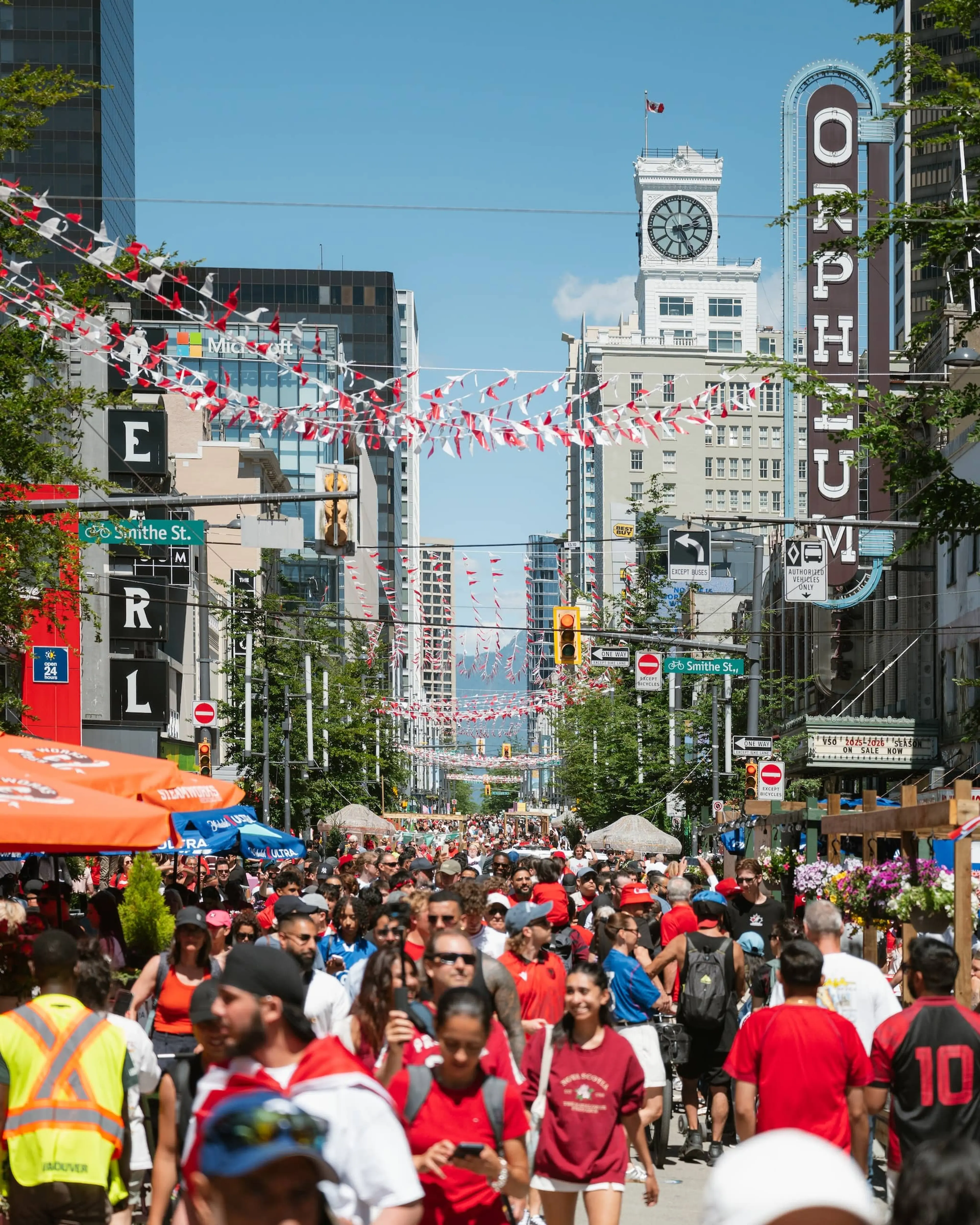 Crowded street festival with people walking under red and white flags, tall buildings, and a clock tower with a Canadian flag.