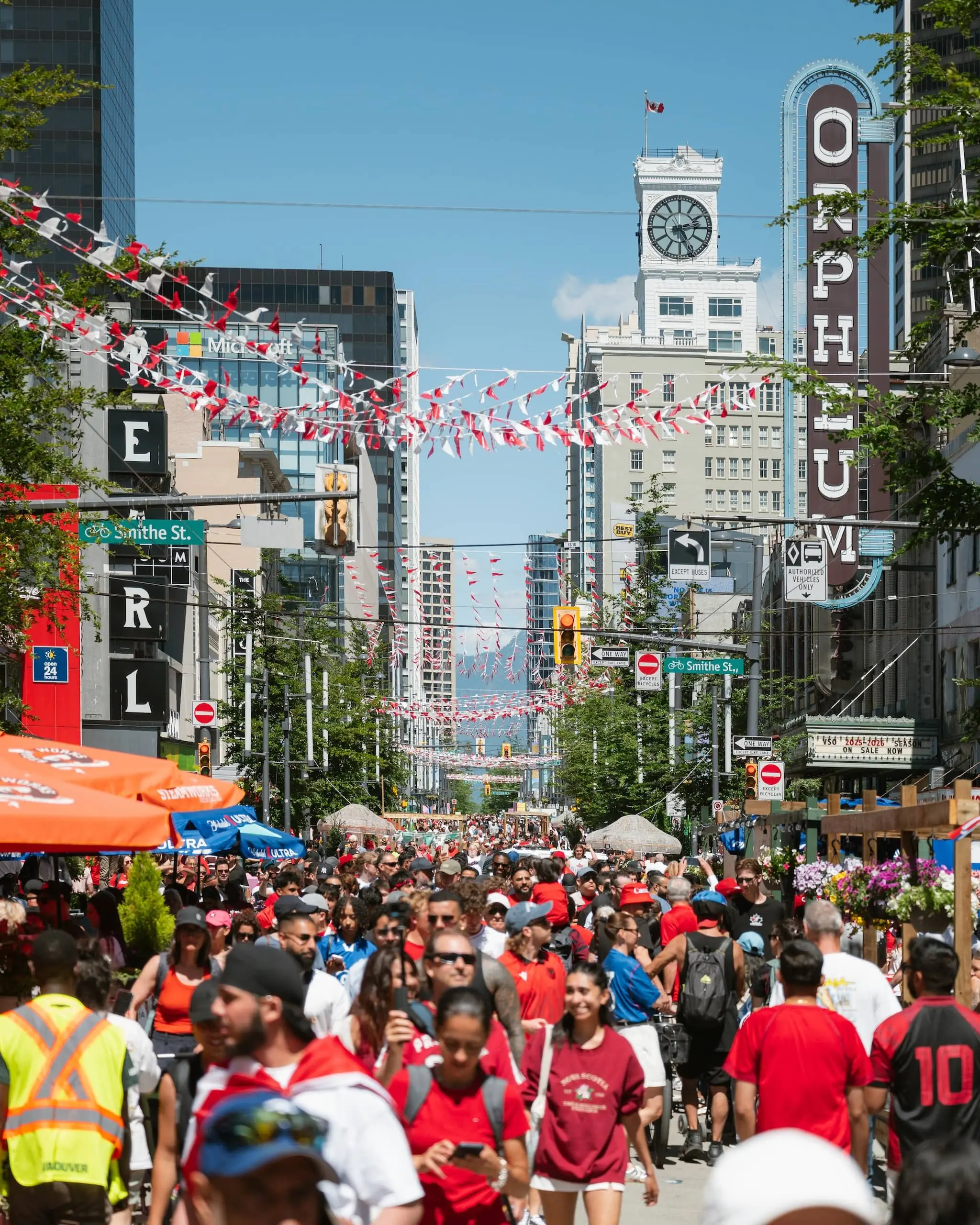 Crowded street festival with people walking under red and white flags, tall buildings, and a clock tower with a Canadian flag.