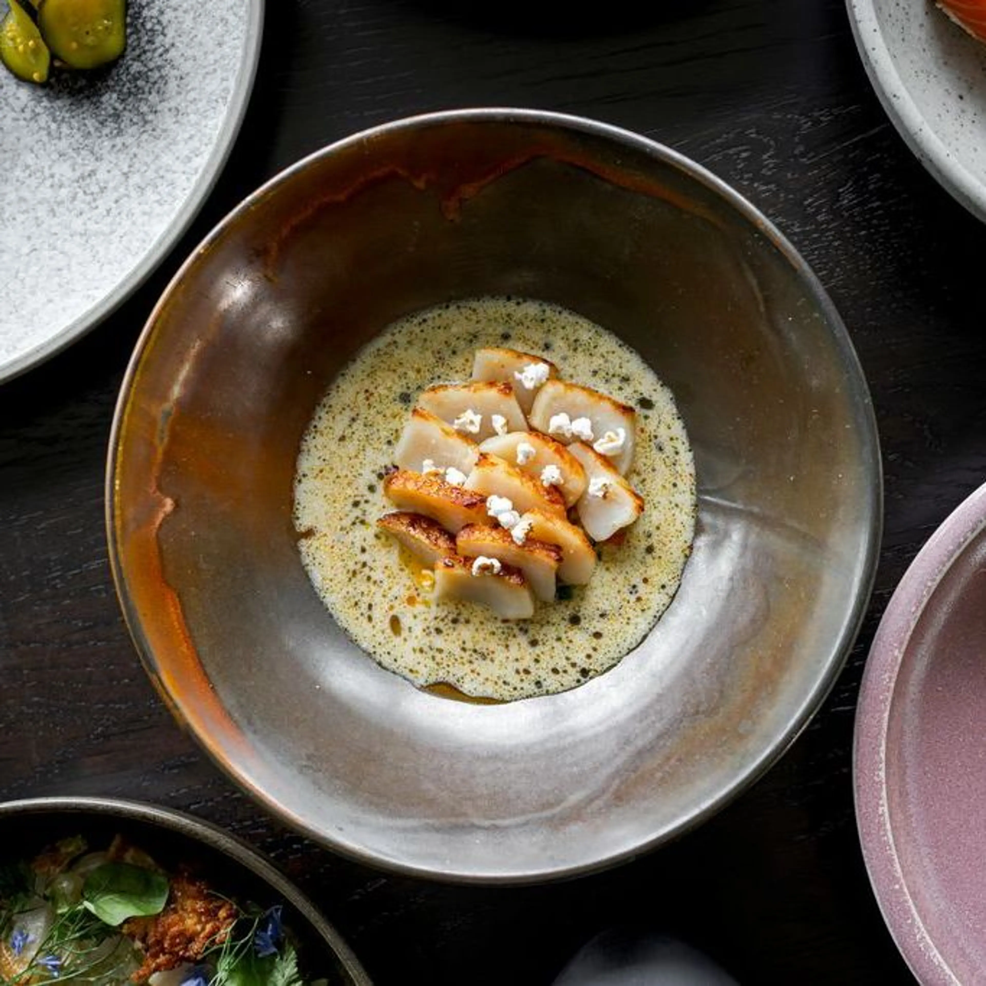 Seared scallops arranged in a circle with foam sauce and small white garnish in a brown ceramic bowl on a dark table.