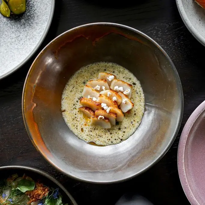 Seared scallops arranged in a circle with foam sauce and small white garnish in a brown ceramic bowl on a dark table.