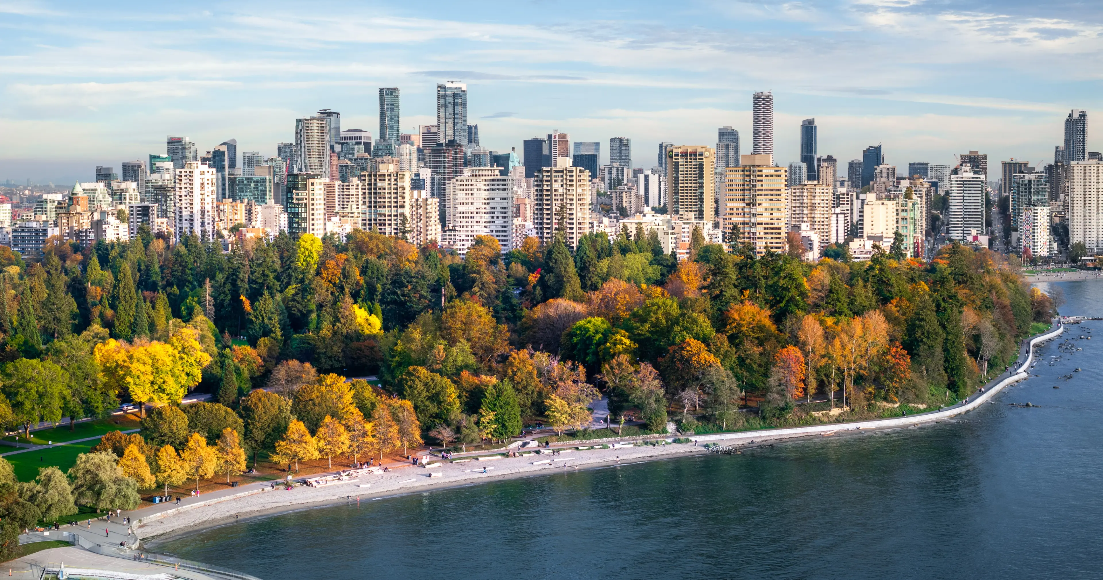 Aerial image of Stanley park with trees in fall colours along the seawall and city skyline in the background.