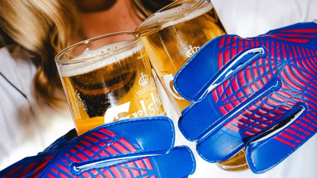 Two hands wearing blue and red gloves clinking glasses of beer with a person in the background.