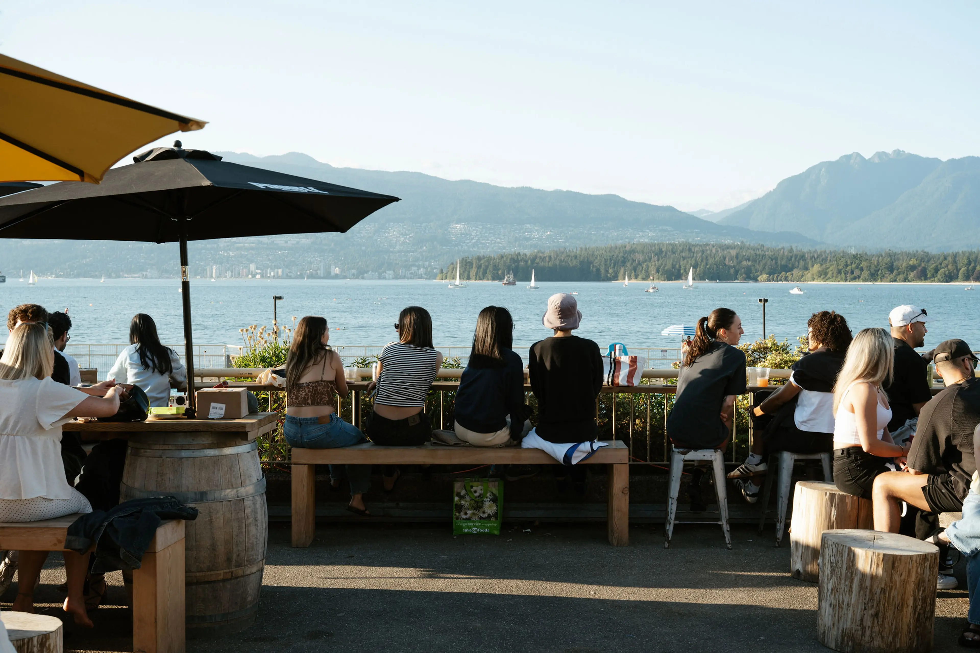 People sitting on benches and stools at an outdoor waterfront cafe with mountains and sailboats in the background.