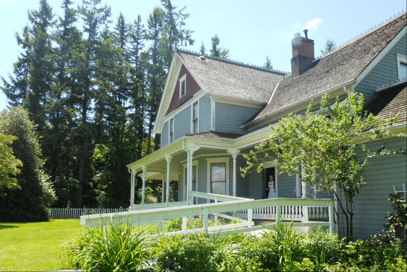 Blue wooden house with a porch and white railing surrounded by green plants and tall trees under a clear sky.