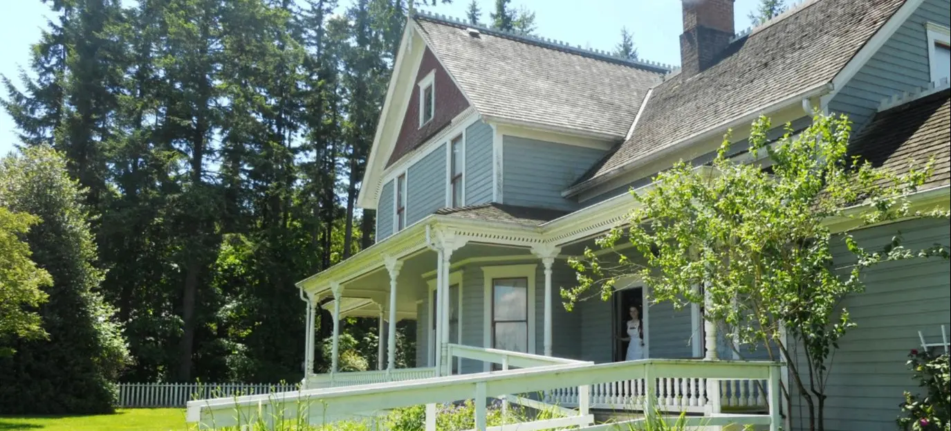 Blue wooden house with a porch and white railing surrounded by green plants and tall trees under a clear sky.