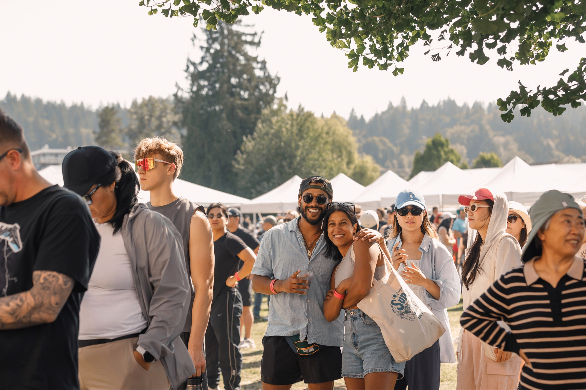 A group of people outdoors at a festival with tents and trees in the background, some smiling and posing for the camera.