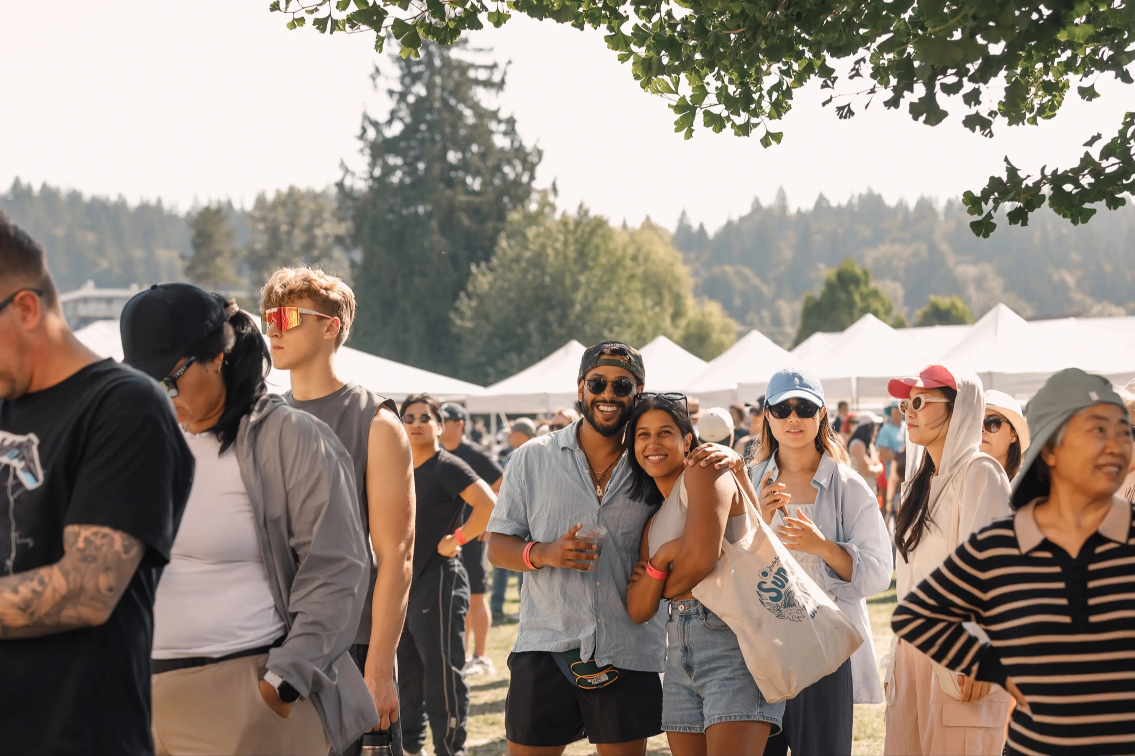 A group of people outdoors at a festival with tents and trees in the background, some smiling and posing for the camera.