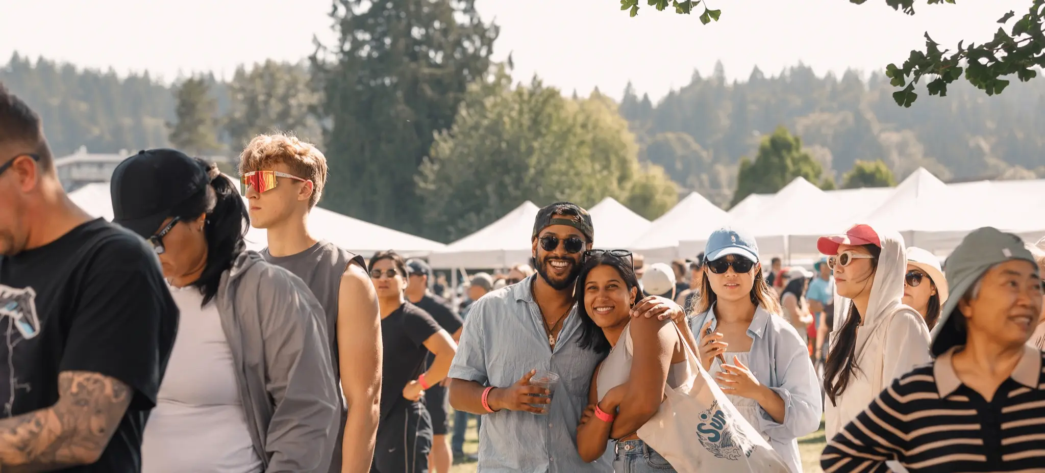 A group of people outdoors at a festival with tents and trees in the background, some smiling and posing for the camera.