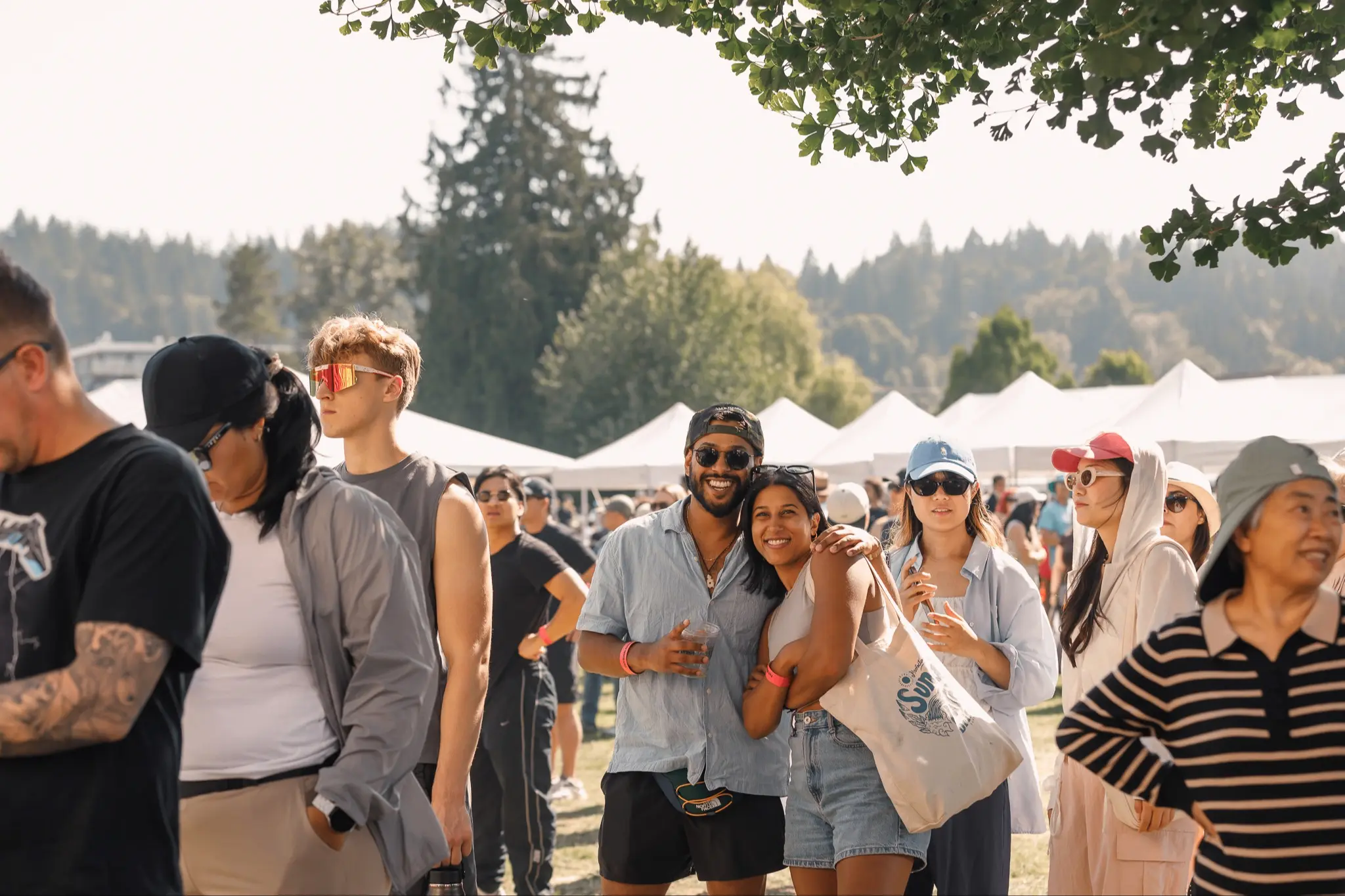 A group of people outdoors at a festival with tents and trees in the background, some smiling and posing for the camera.