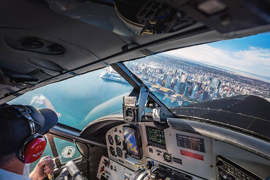 Pilot wearing headset flying a plane over a coastal city with water and buildings visible through the cockpit window.