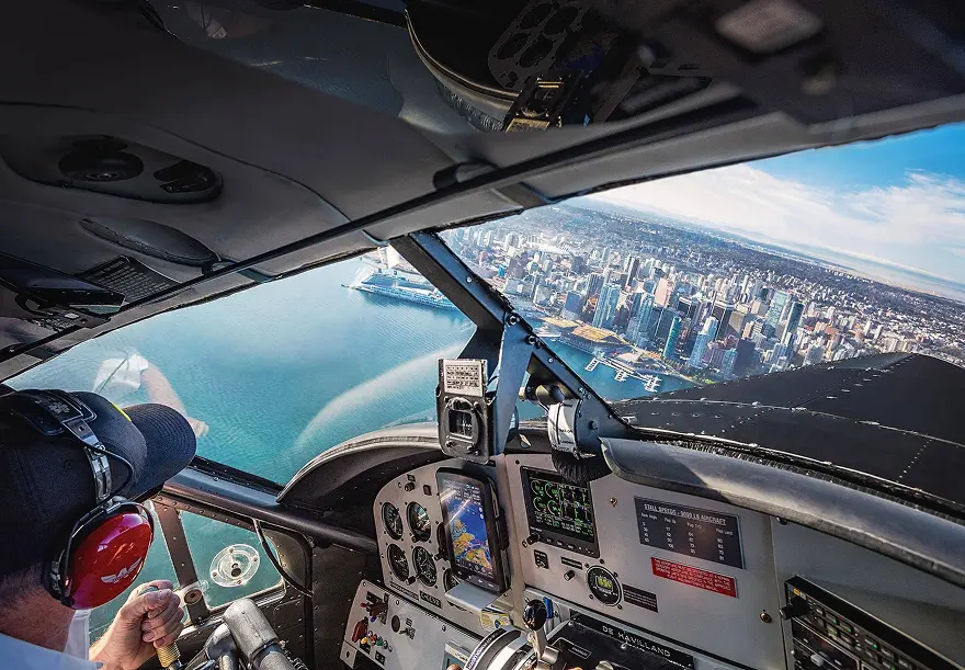 Pilot wearing headset flying a plane over a coastal city with water and buildings visible through the cockpit window.