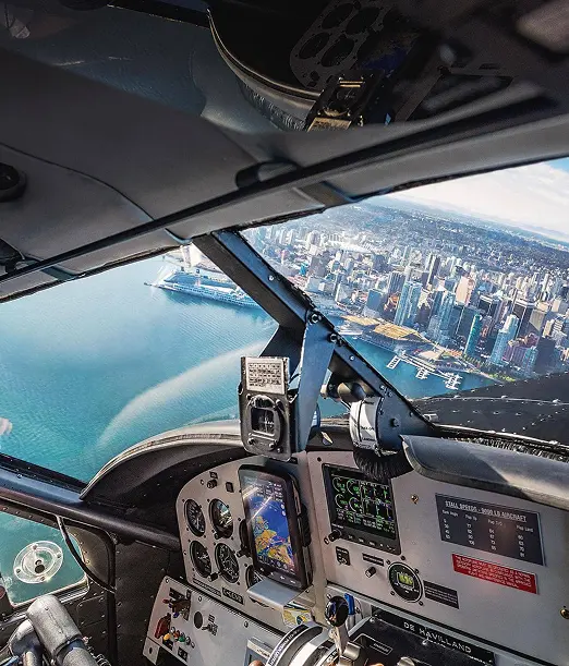 Pilot wearing headset flying a plane over a coastal city with water and buildings visible through the cockpit window.