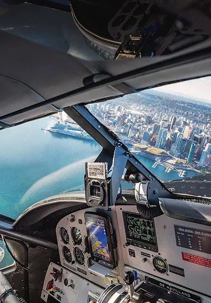 Pilot wearing headset flying a plane over a coastal city with water and buildings visible through the cockpit window.