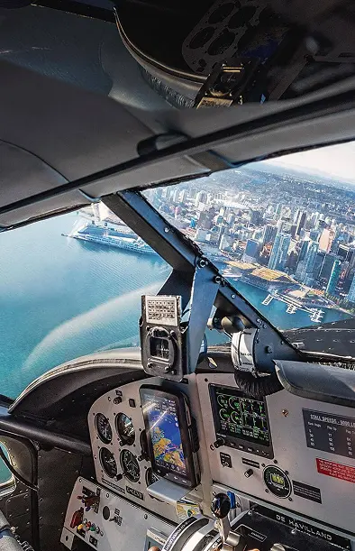 Pilot wearing headset flying a plane over a coastal city with water and buildings visible through the cockpit window.
