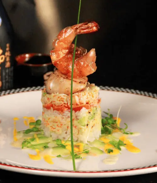 Gourmet dish with layered rice and vegetables topped with grilled shrimp, garnished with green herbs and yellow sauce on a white plate.