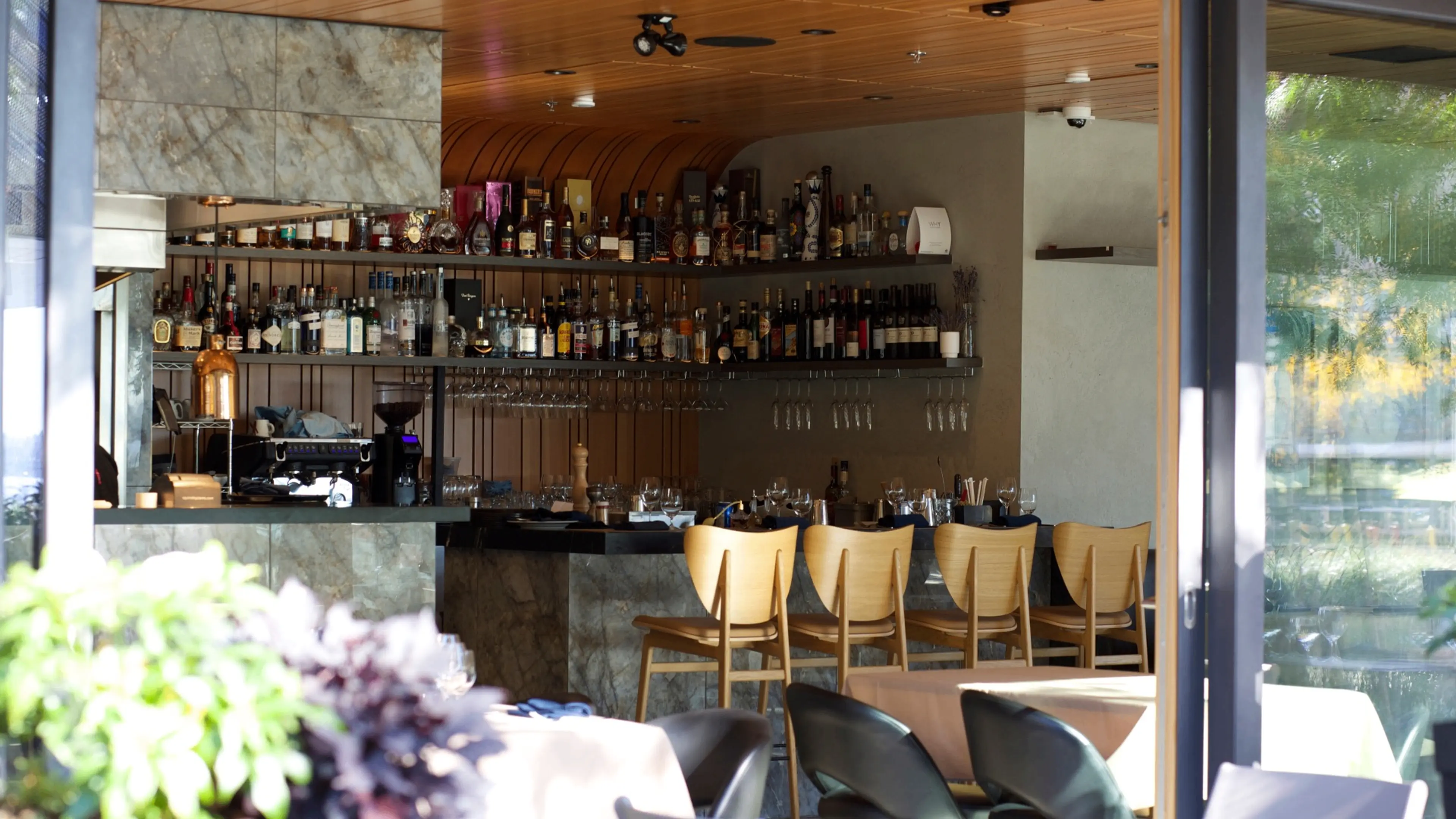 Empty bar area with wooden chairs, shelves of liquor bottles, and glassware in a modern restaurant.