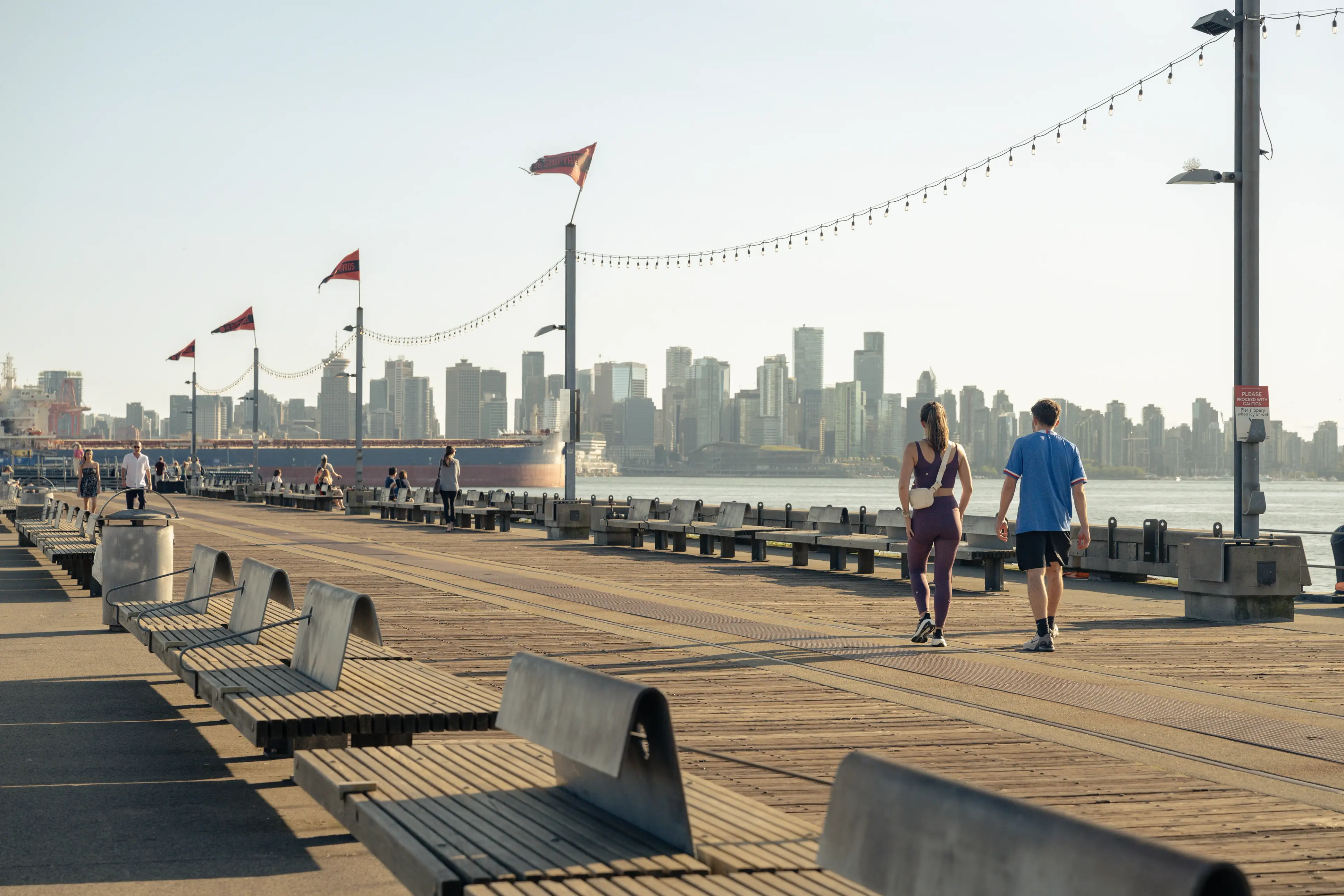 Two people walk along the pier at the Shipyards District with the downtown Vancouver skyline in the background.