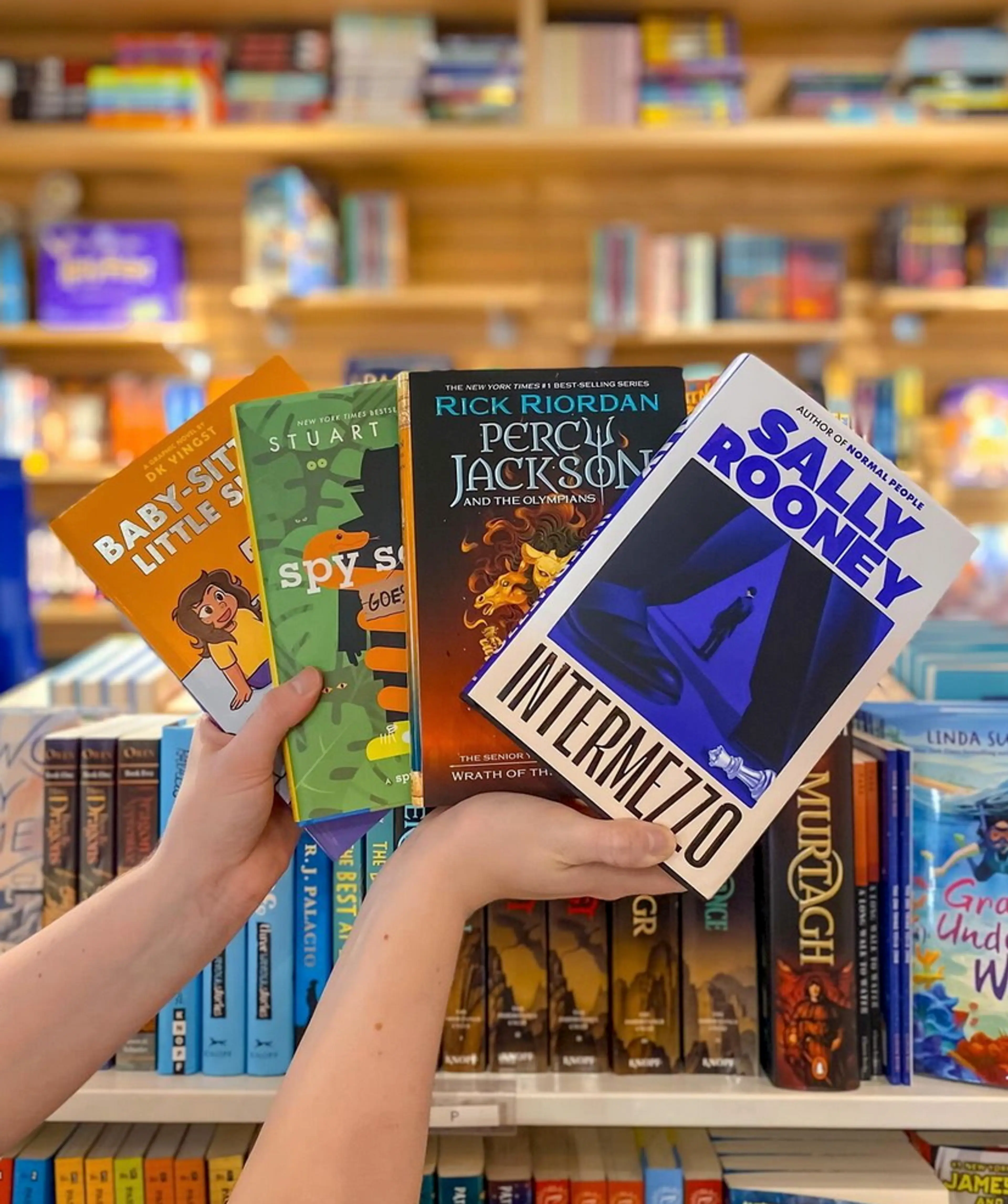 Two hands hold up four young adult books. There are shelves of more books in the background.