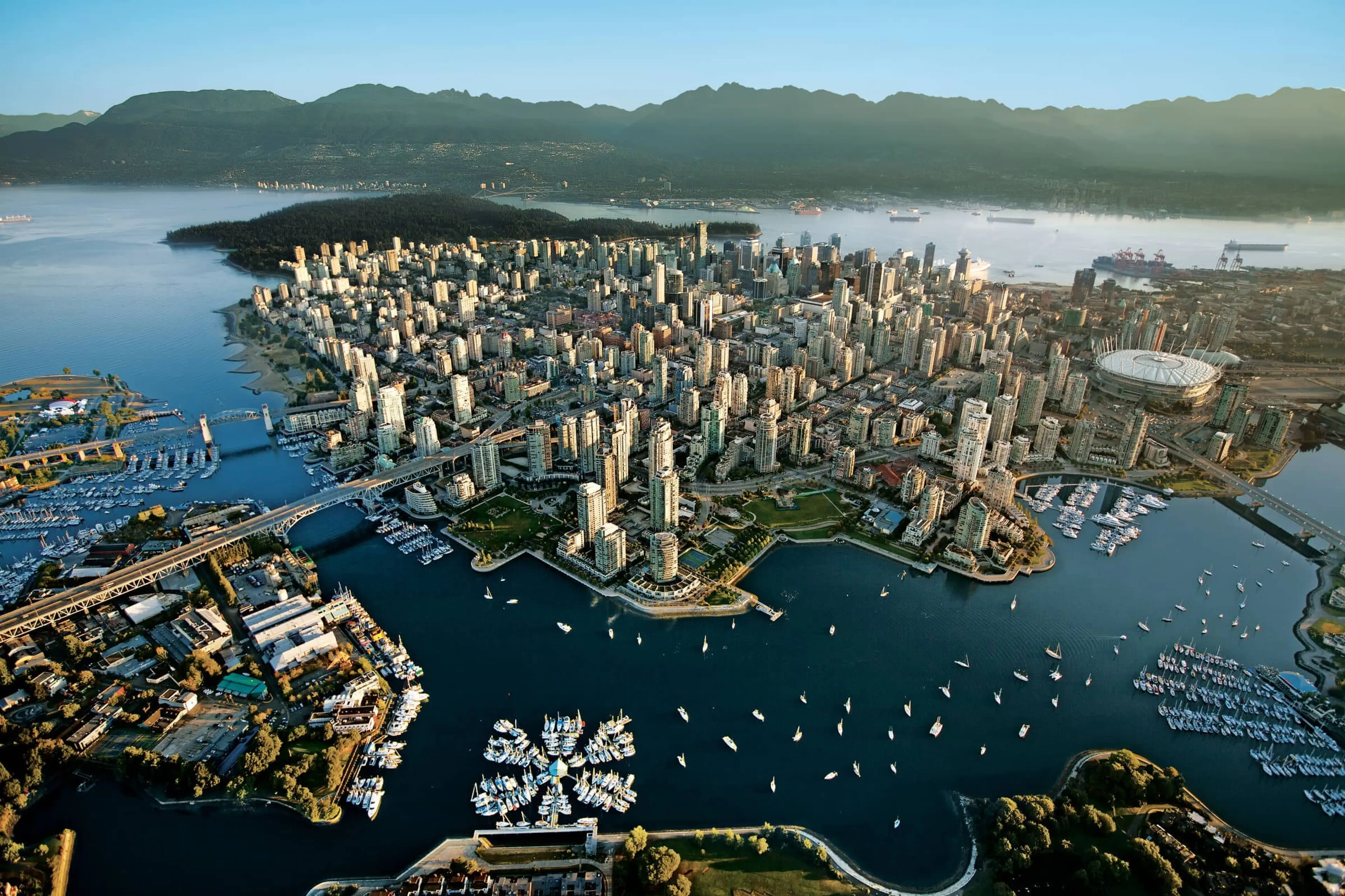 Aerial view of a coastal city with numerous high-rise buildings, bridges, marinas with boats, and mountains in the background.