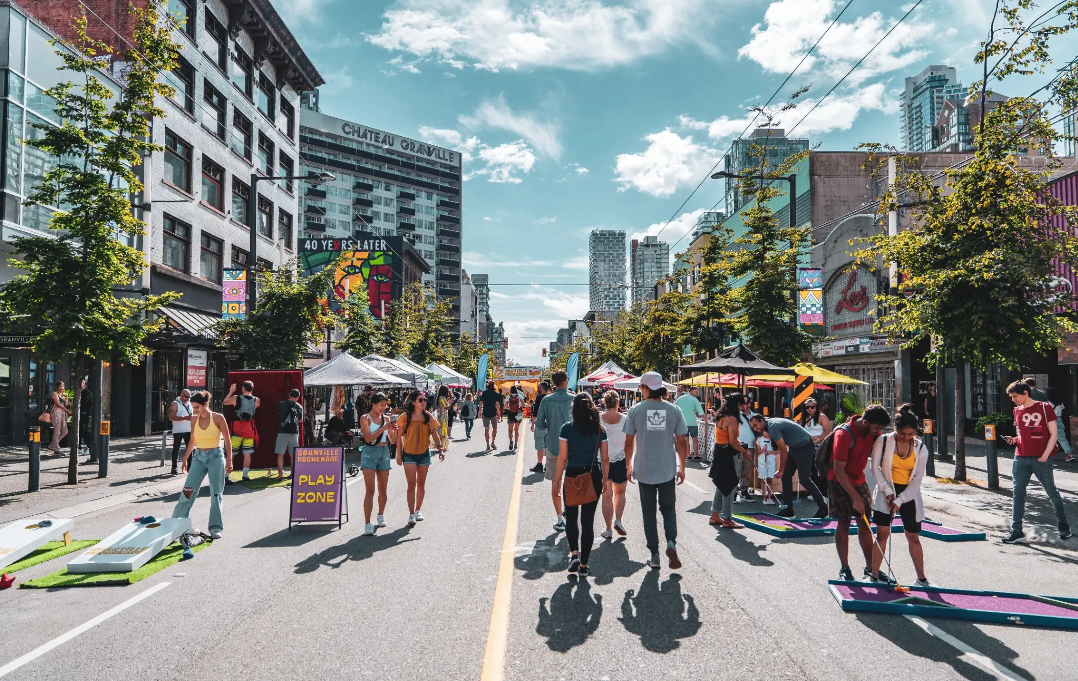 People walking and playing mini golf on a sunny city street with tents and buildings on both sides.