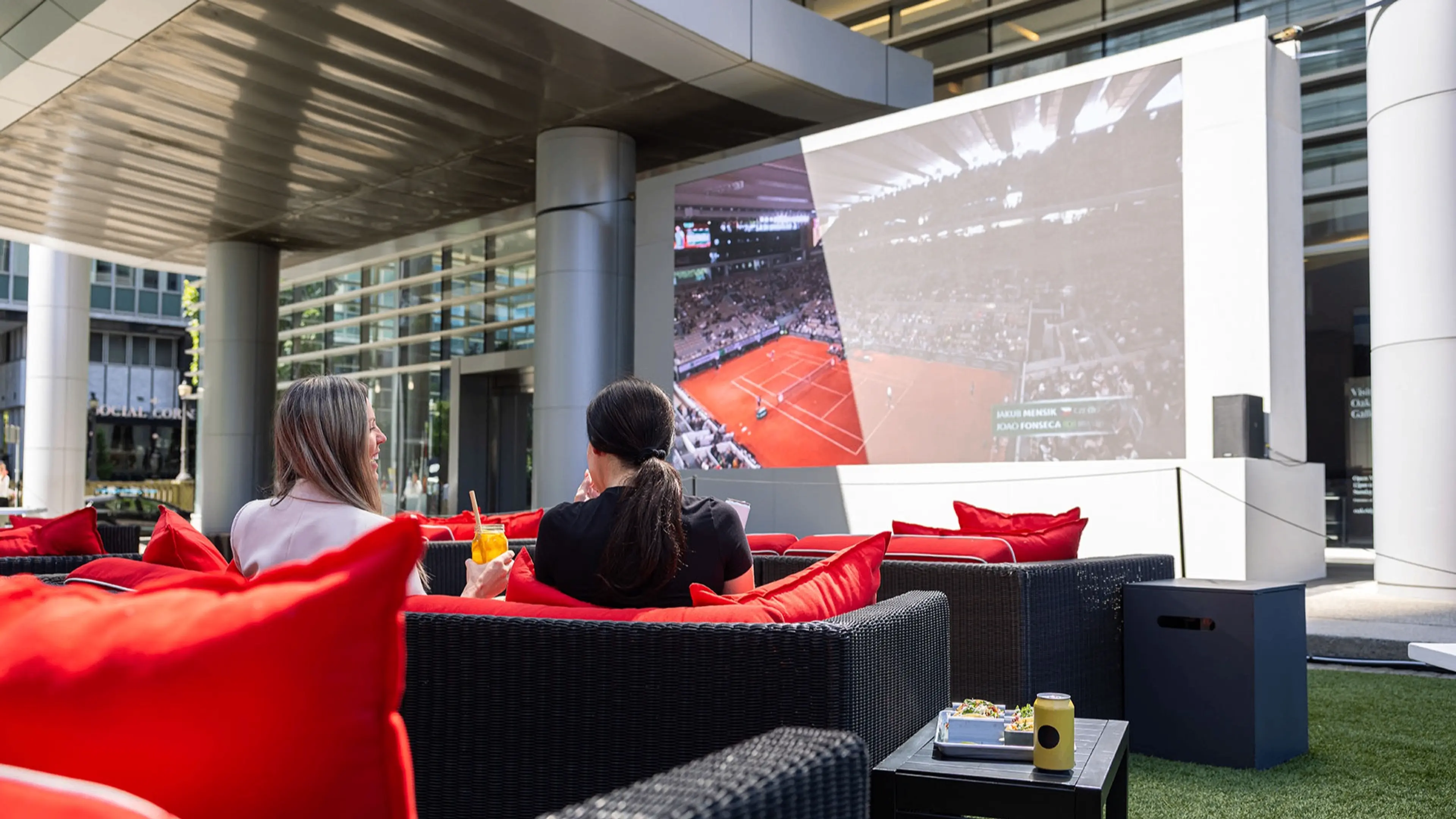 Two people sitting on outdoor black wicker sofas with red cushions watching a tennis match on a large screen.