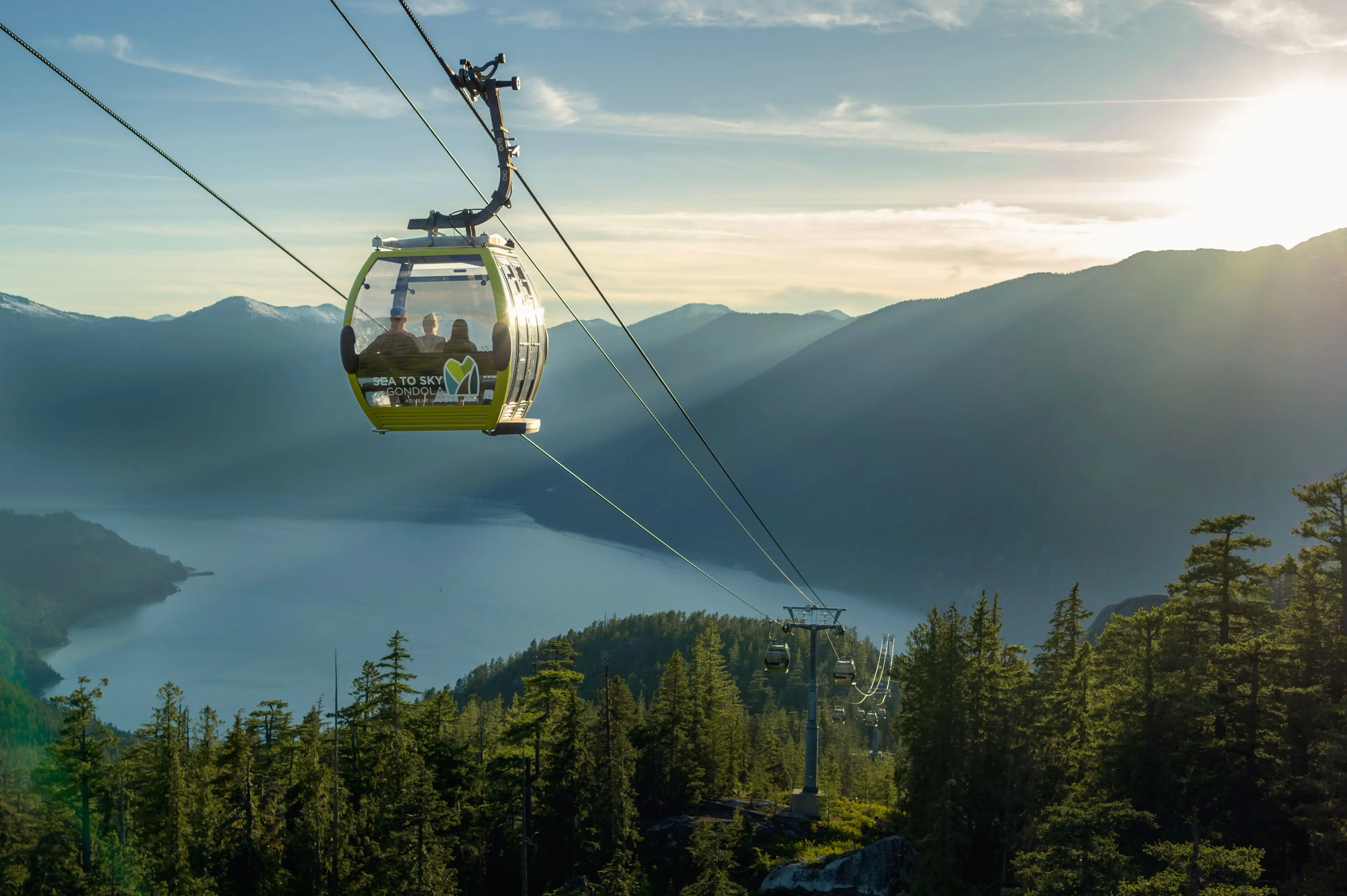 Sea to Sky Gondola lift with passengers over forested mountains and a lake at sunset.