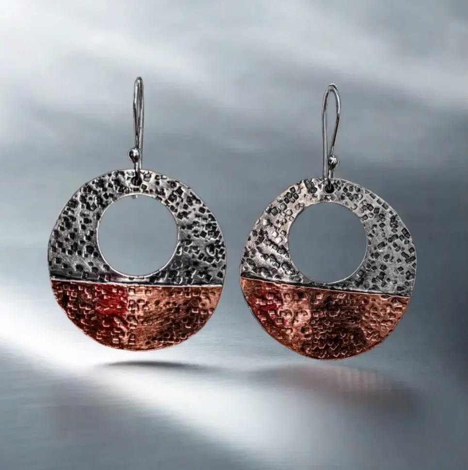 A pair of hand-made silver and copper circular earrings set against a grey background.