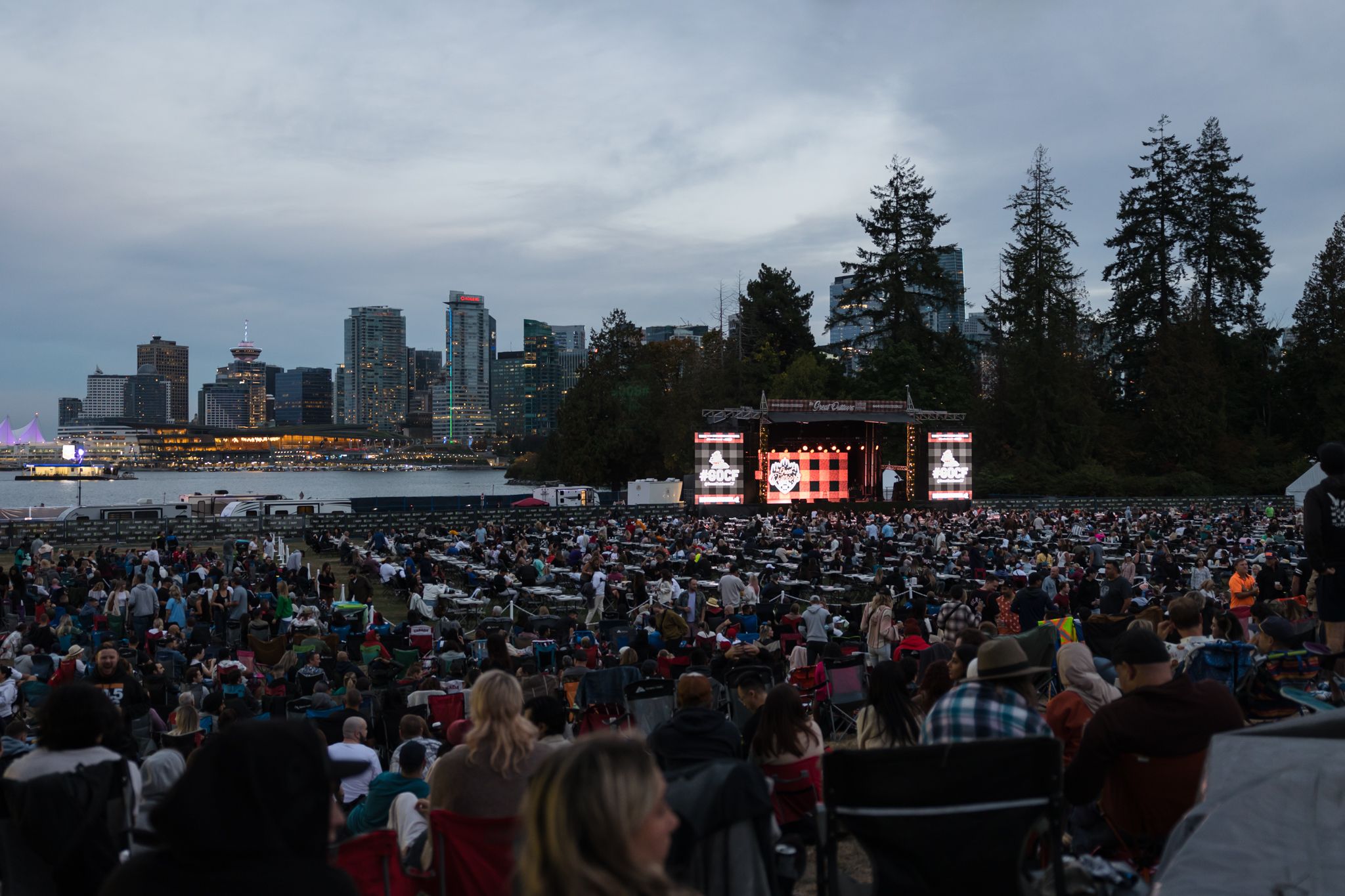 Large outdoor crowd seated facing a stage with city skyline and water in the background at dusk.