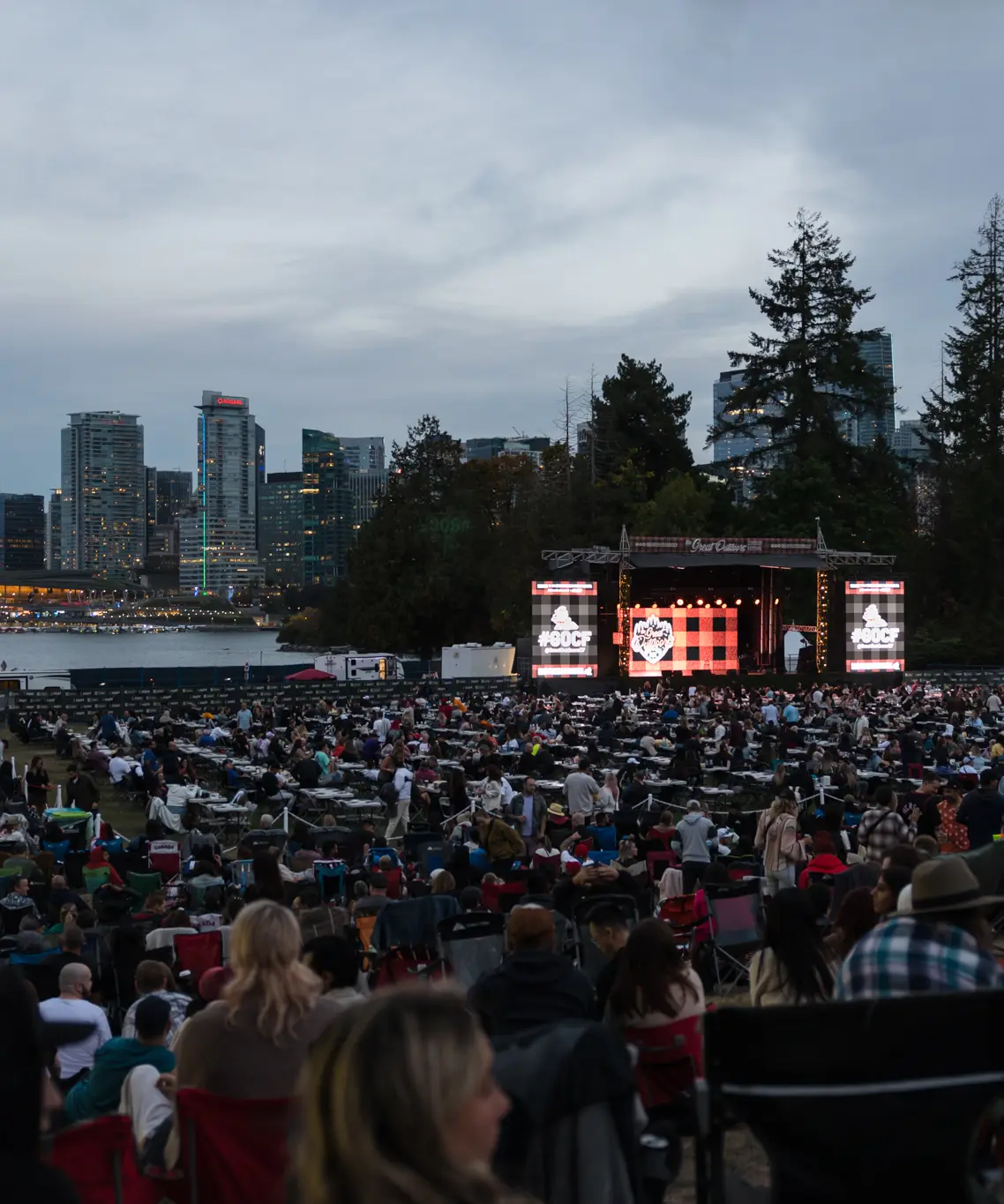 Large outdoor crowd seated facing a stage with city skyline and water in the background at dusk.